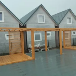 Drewniana konstrukcja z elementami ażurowymi na szarym tarasie kompozytowym, tło stanowią identyczne domki w jasnej kolorystyce, widoczne ślady deszczu.