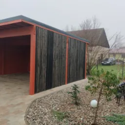 Zadaszone miejsce parkingowe z drewnianą konstrukcją pomalowaną na czerwono, z czarnymi pionowymi deskami na ścianie bocznej, wyłożone kostką brukową, z ozdobnym żwirowym obrzeżem i małym drzewkiem...