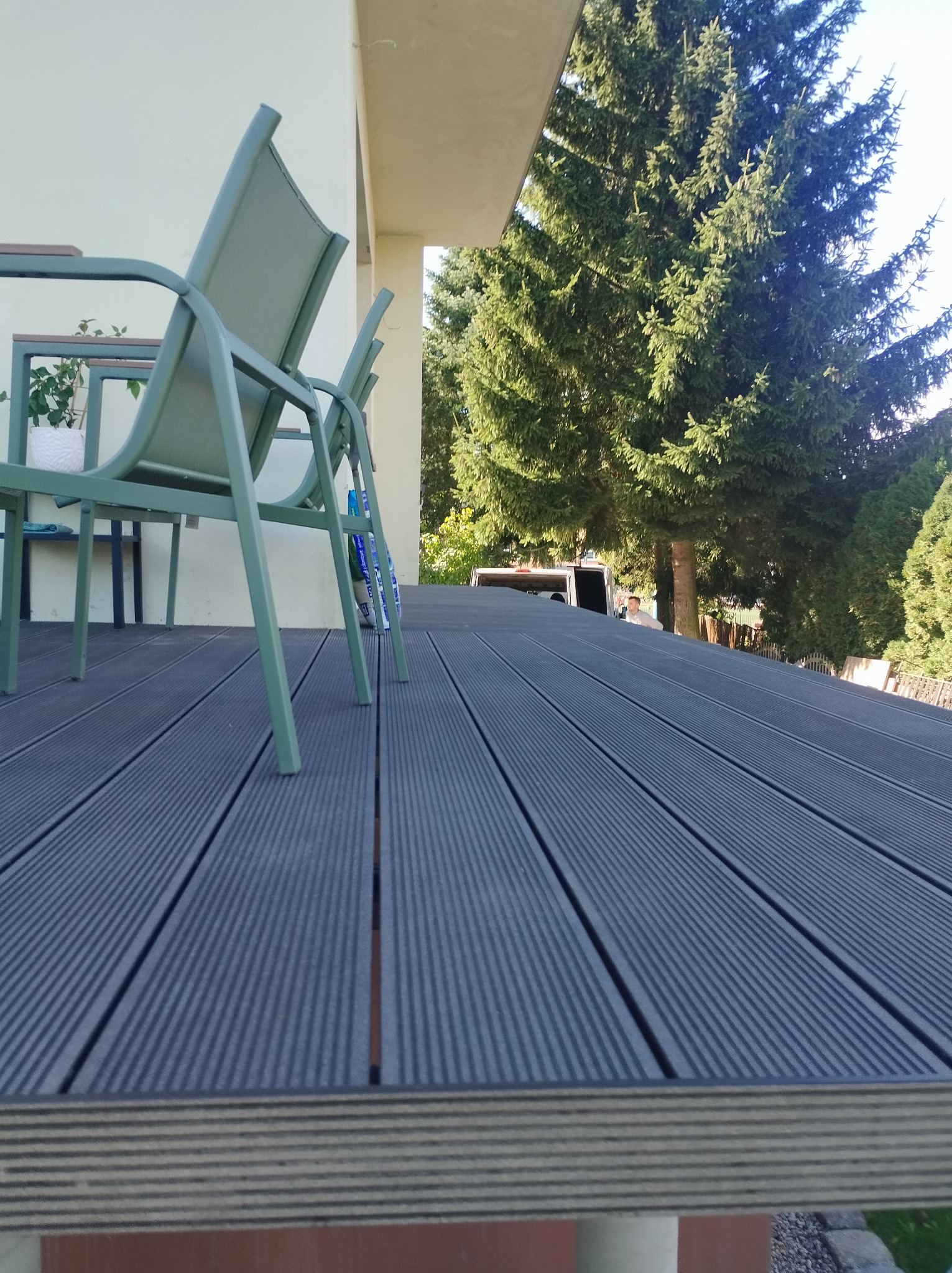 Nowoczesny taras z desek kompozytowych w odcieniach szarości, z zielonymi krzesłami ogrodowymi i widokiem na iglaste drzewa w tle.