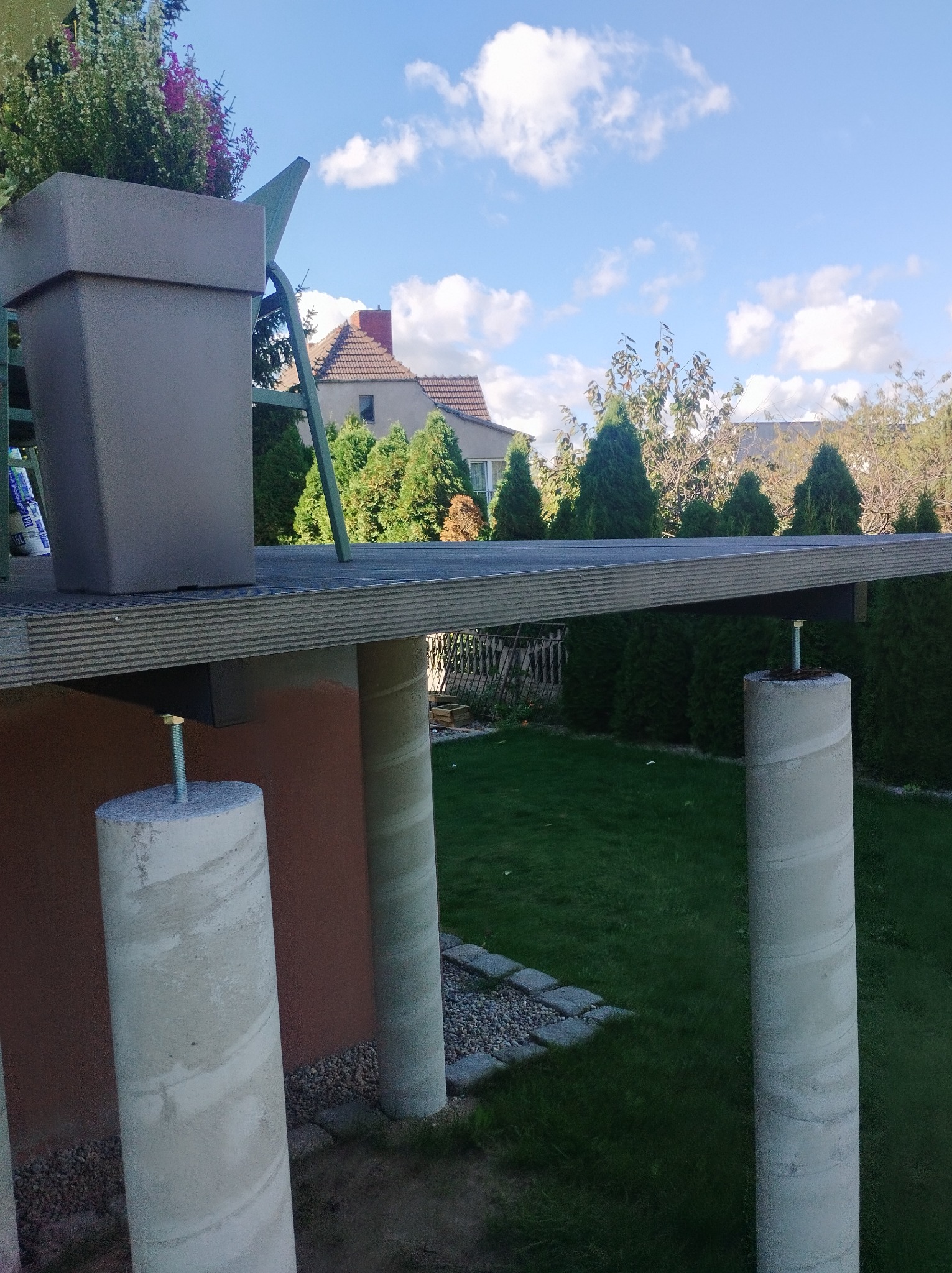 Konstrukcja z betonowymi słupami i drewnianym blatem, widoczna z boku, z donicą na górze, na tle zielonego ogrodu i błękitnego nieba z chmurami.