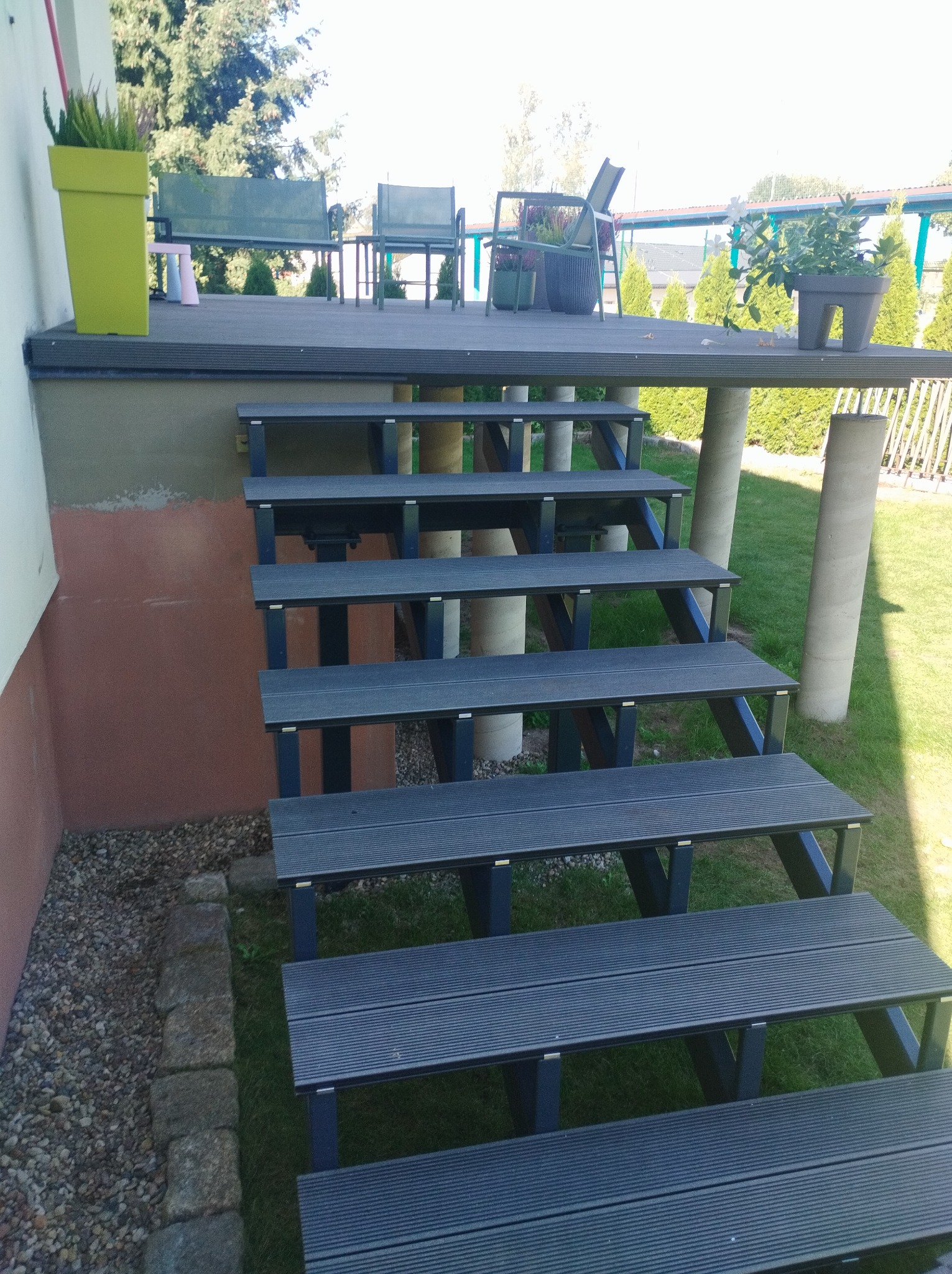 Schody z desek kompozytowych prowadzące na podwyższony taras z meblami ogrodowymi i roślinami w donicach, widok z boku na tle trawnika i drzew