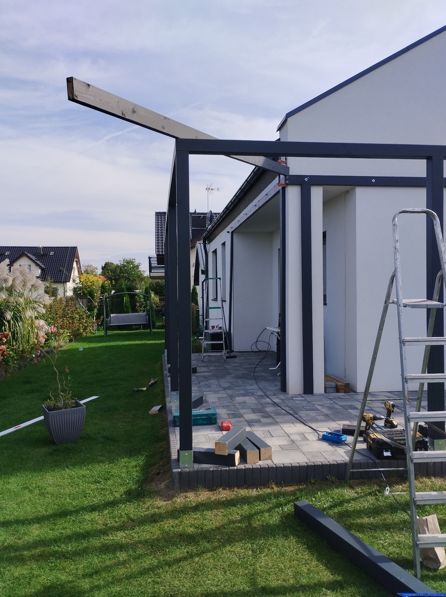 Montaż metalowej konstrukcji z elementami drewnianymi, prawdopodobnie pergoli, widoczny na tle domu z ogrodem, z narzędziami i materiałami budowlanymi w pobliżu.