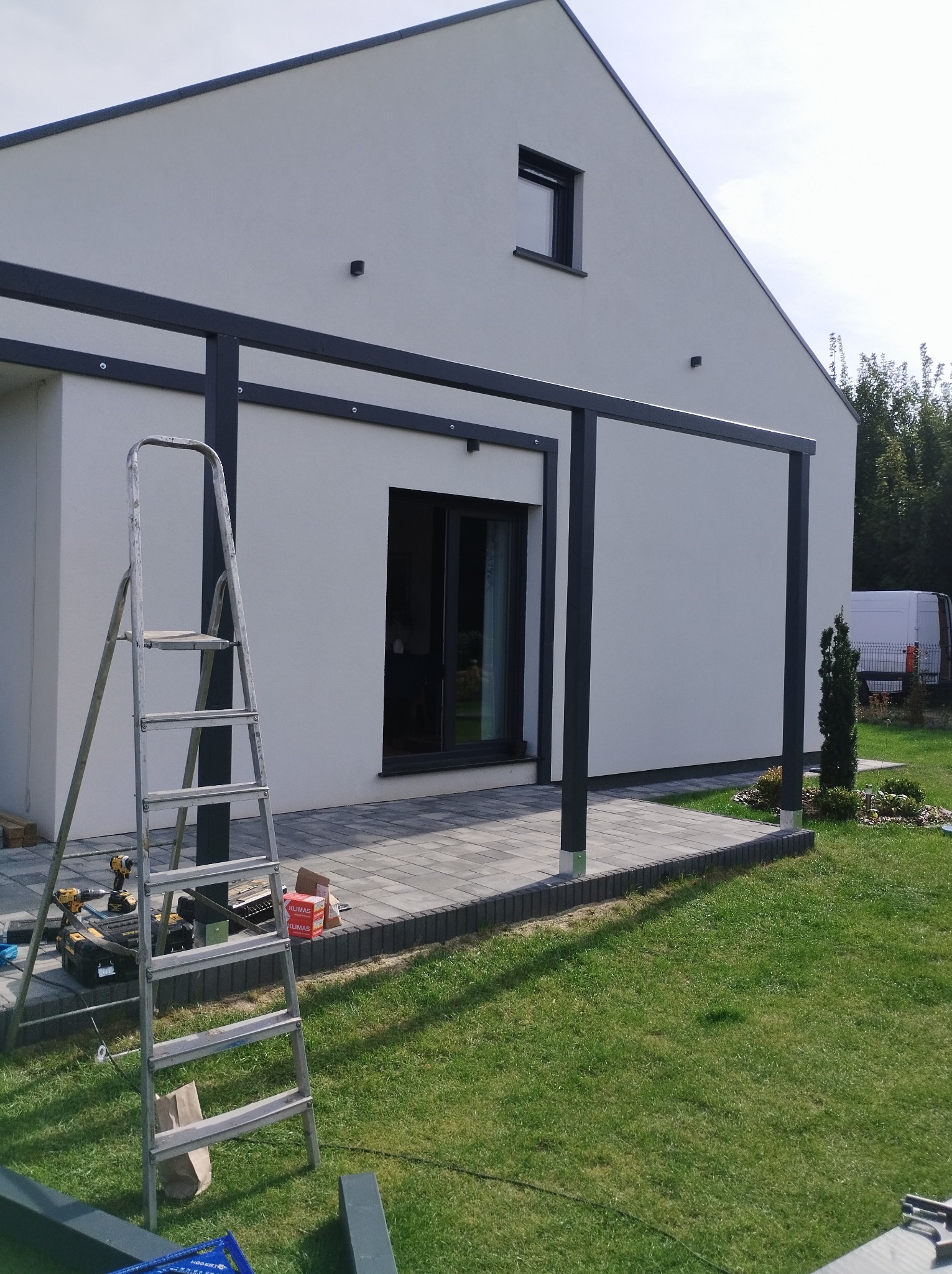 Montaż metalowej konstrukcji pergoli przy nowoczesnym domu; widoczna drabina, narzędzia i kostka brukowa na trawniku.