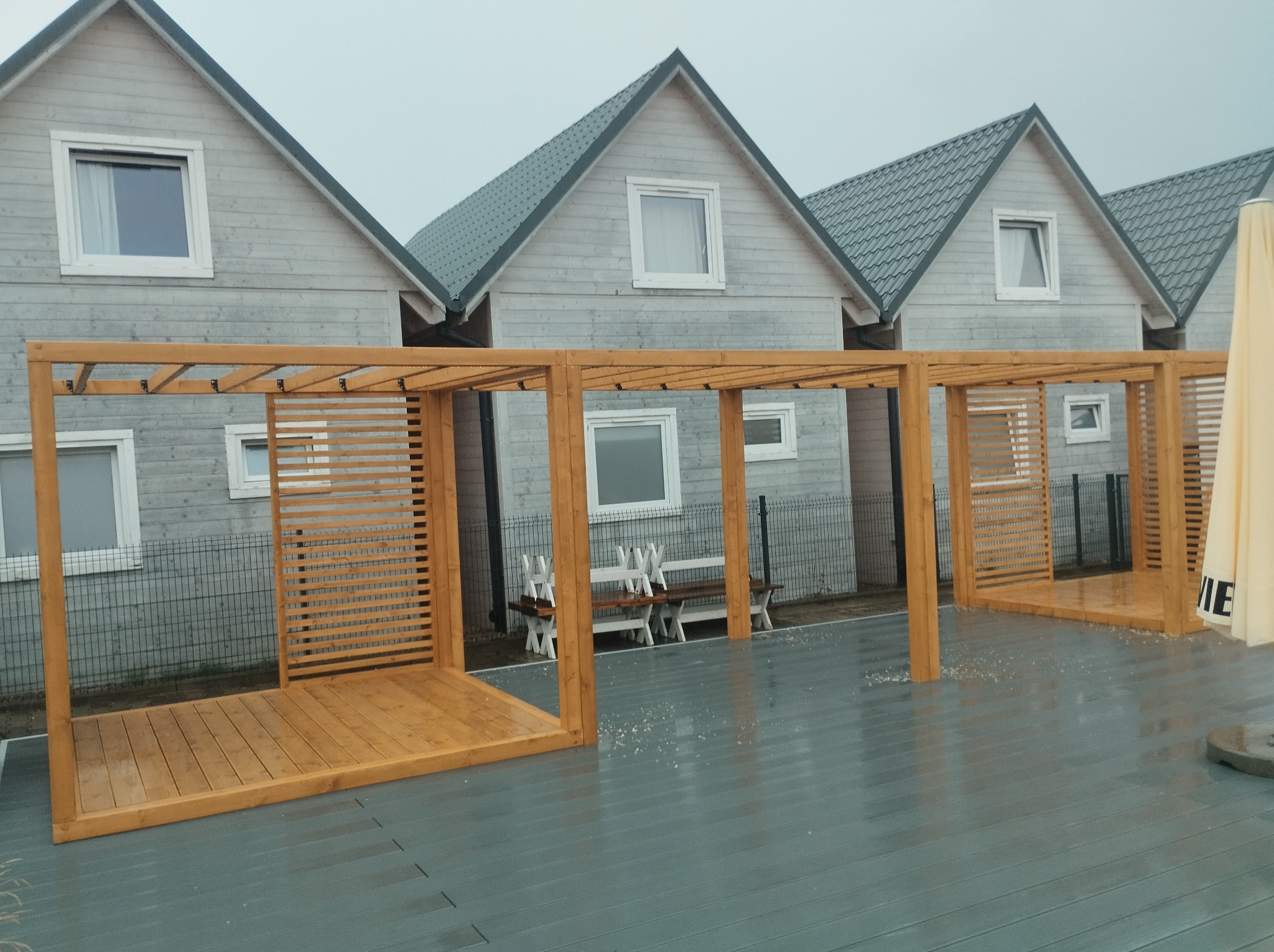 Drewniana konstrukcja z elementami ażurowymi na szarym tarasie kompozytowym, tło stanowią identyczne domki w jasnej kolorystyce, widoczne ślady deszczu.