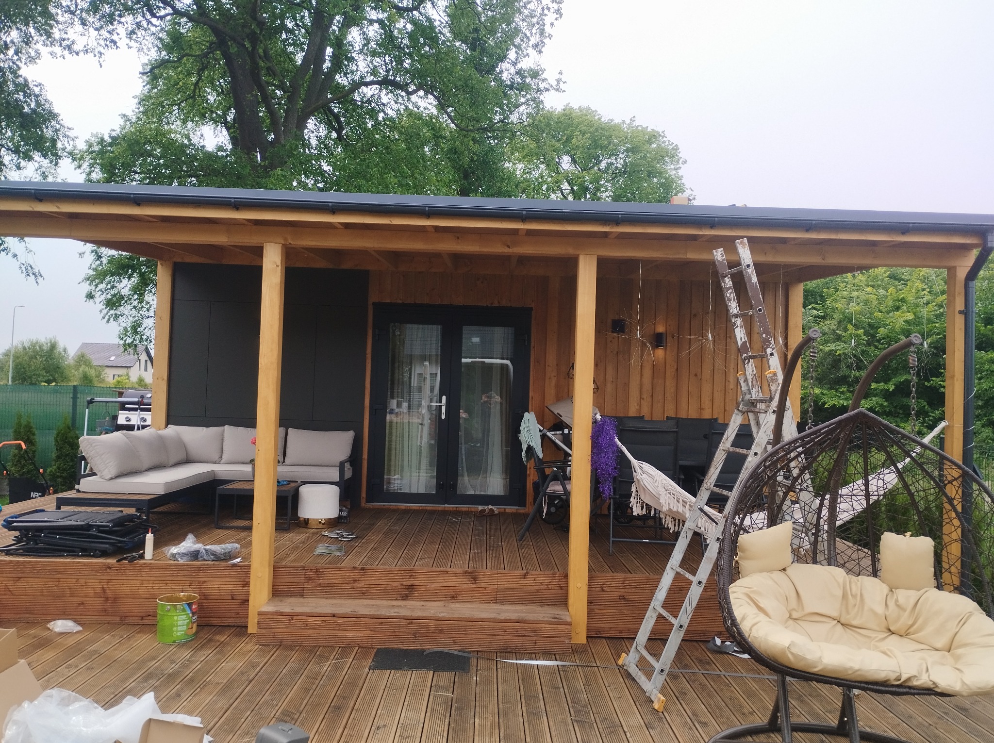 Zadaszony taras drewniany w trakcie wykańczania, widoczna drabina, farba, meble ogrodowe i huśtawka wisząca