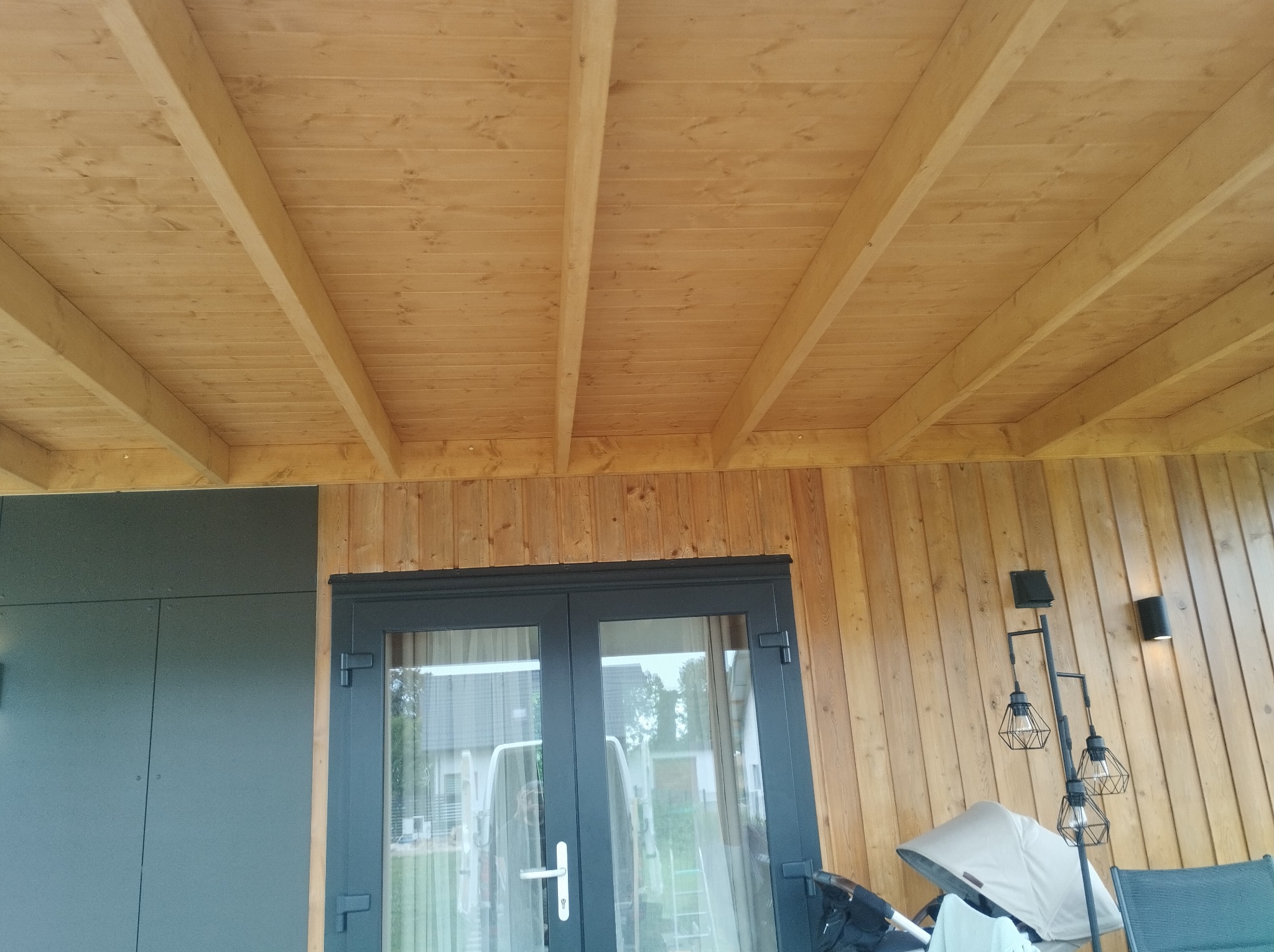 Drewniany sufit z belkami w zadaszonej przestrzeni, widok z dołu, fragment ściany z pionowych desek i przeszklone drzwi w czarnej ramie.