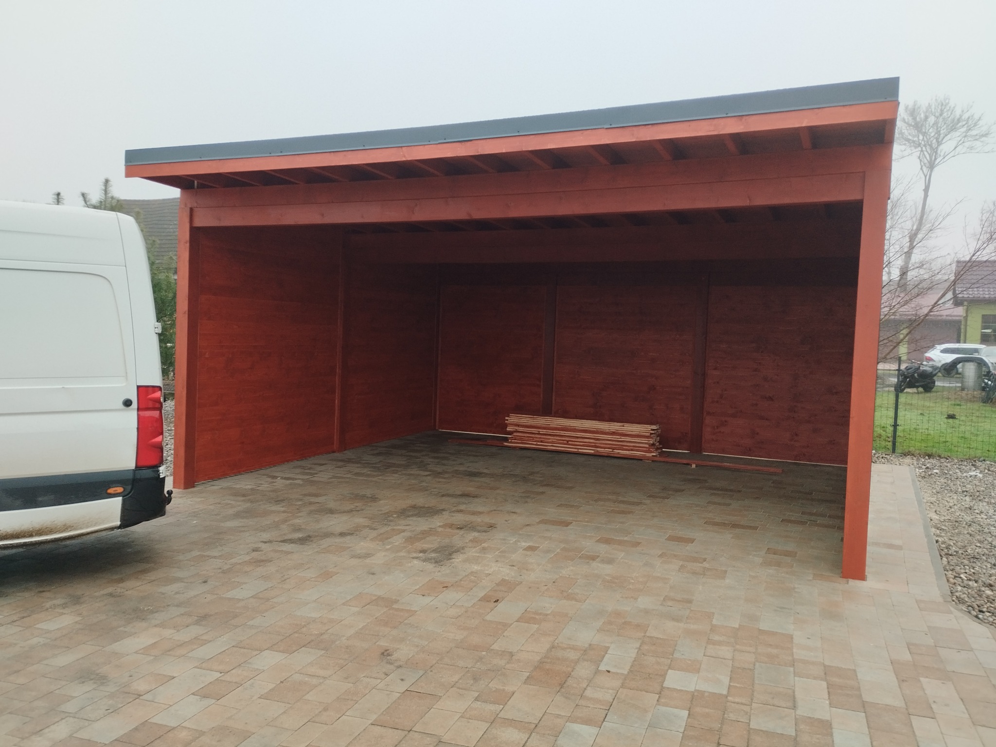 Ciemnobrązowa, drewniana wiata garażowa z szarym dachem na kostce brukowej, częściowo zasłonięta białym samochodem dostawczym, z ułożonymi deskami w środku, w tle domy i drzewa we mgle.