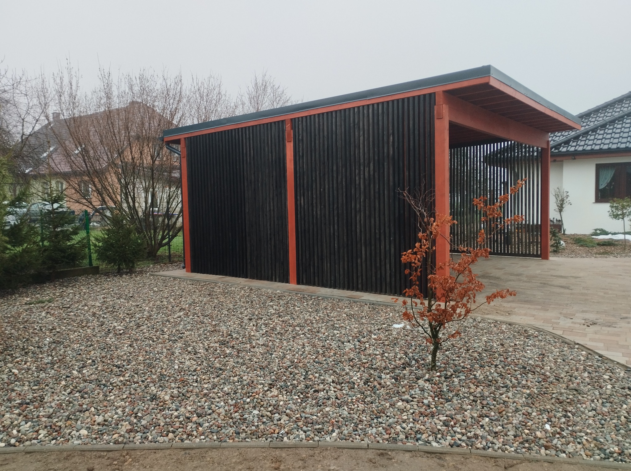 Nowoczesna konstrukcja z ciemnego drewna z pomarańczowymi elementami, prawdopodobnie zadaszenie na samochód, otoczona kamieniami i małym drzewkiem z brązowymi liśćmi na tle pochmurnego nieba.