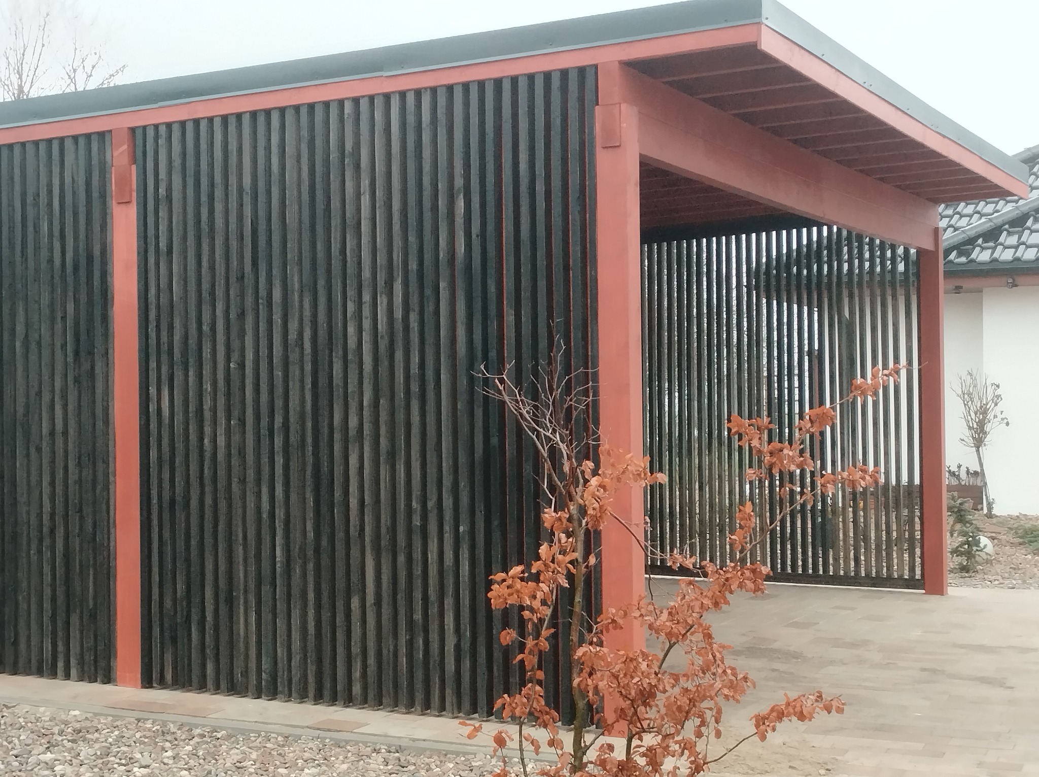 Nowoczesna wiata garażowa z ciemnymi, pionowymi deskami i pomarańczowymi elementami konstrukcyjnymi, widoczna z zewnątrz, z małym drzewkiem na pierwszym planie.