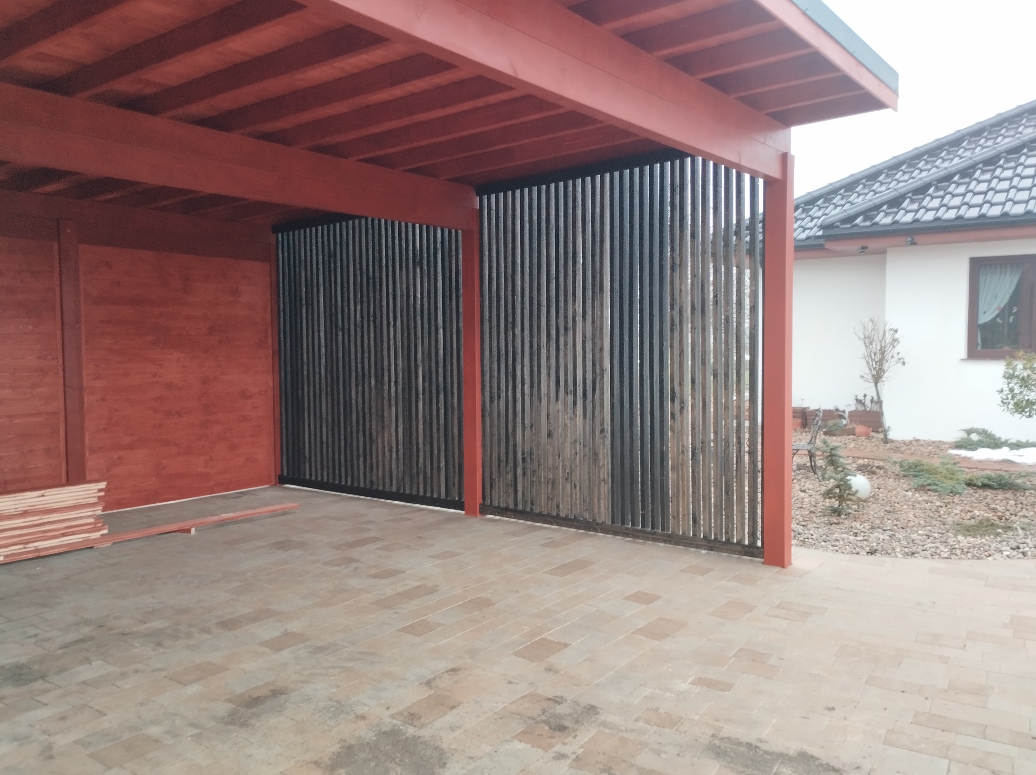 Zadaszone miejsce parkingowe z drewnianą konstrukcją w kolorze ceglastym i pionowymi, grafitowymi panelami z drewna, wyłożone ceglastą kostką brukową, z widocznym stosem desek i fragmentem ogrodu...