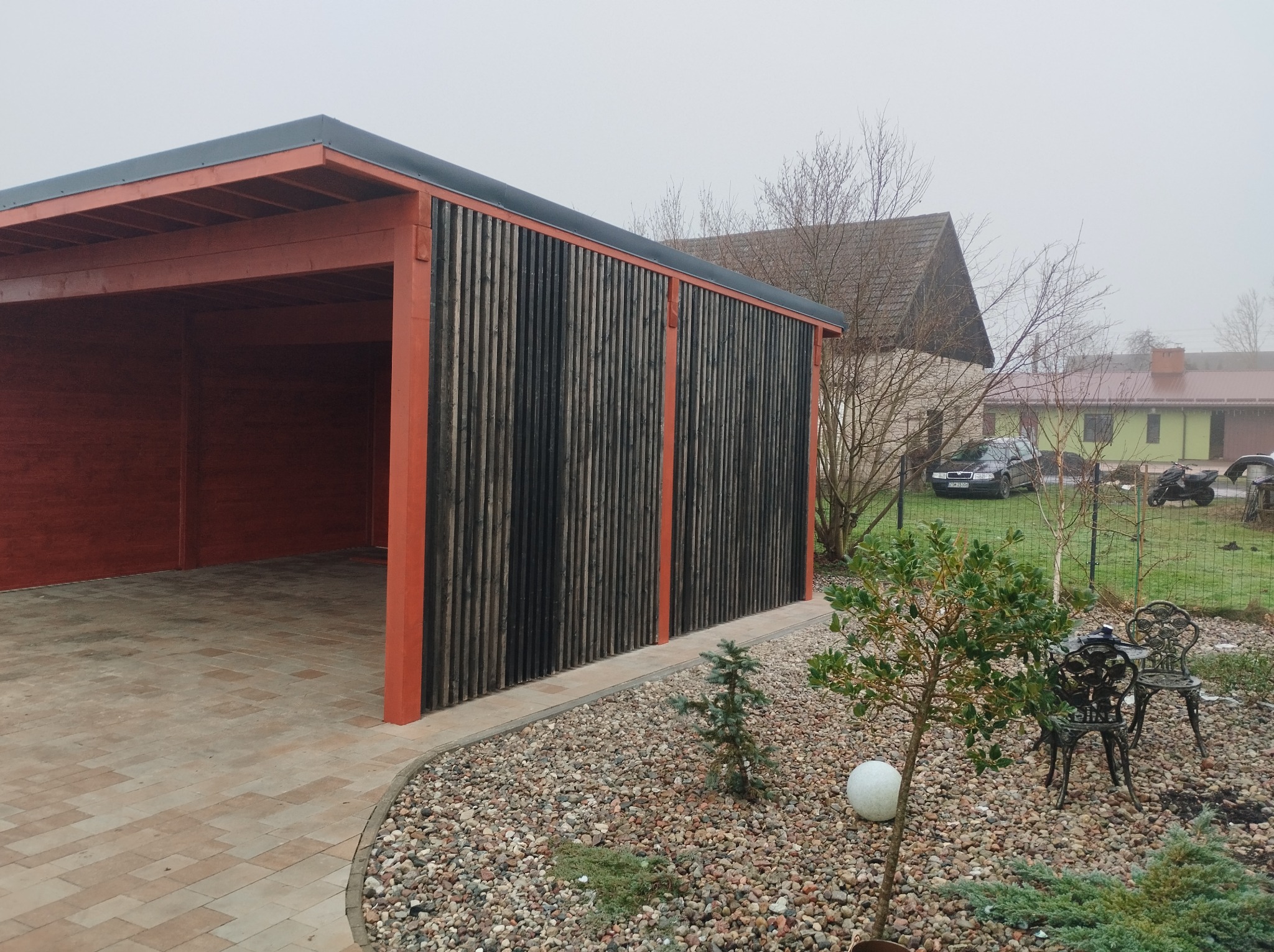Zadaszone miejsce parkingowe z drewnianą konstrukcją pomalowaną na czerwono, z czarnymi pionowymi deskami na ścianie bocznej, wyłożone kostką brukową, z ozdobnym żwirowym obrzeżem i małym drzewkiem...