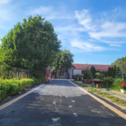 Świeżo ułożona, czarna kostka brukowa z jasnymi obrzeżami prowadząca do domu, widoczne ślady stóp, pracownik zagęszcza powierzchnię, błękitne niebo z chmurami w tle.