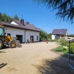 Żółta ładowarka teleskopowa na żwirowym podłożu przed nowoczesnym domem z brązowym dachem, w tle widoczny drugi budynek i osoba spacerująca po żwirowym podwórku.