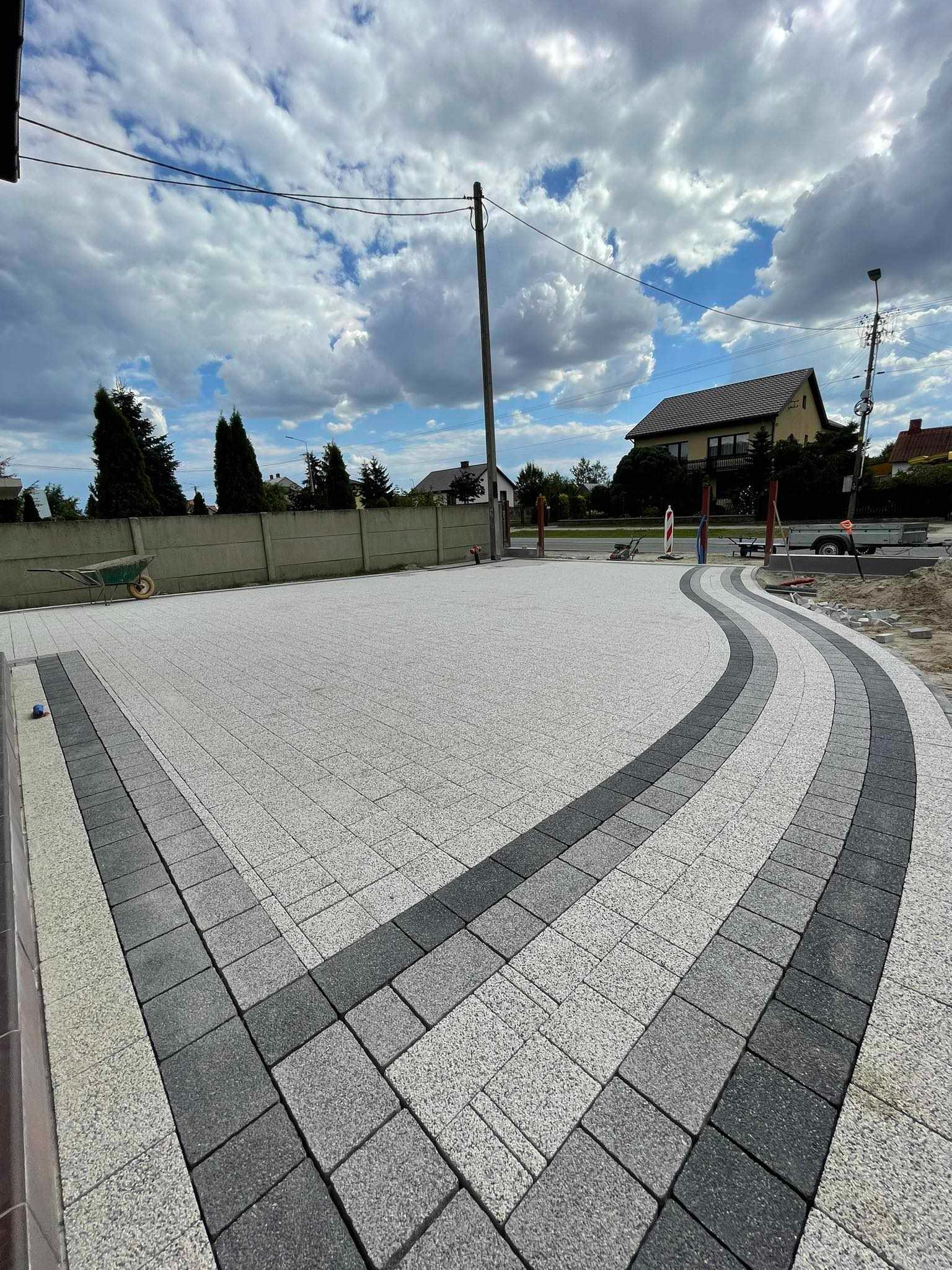 Nowo ułożona, jasnoszara kostka brukowa z ciemnoszarymi obrzeżami, tworząca łukowaty wzór wjazdowy na tle domu i pochmurnego nieba.