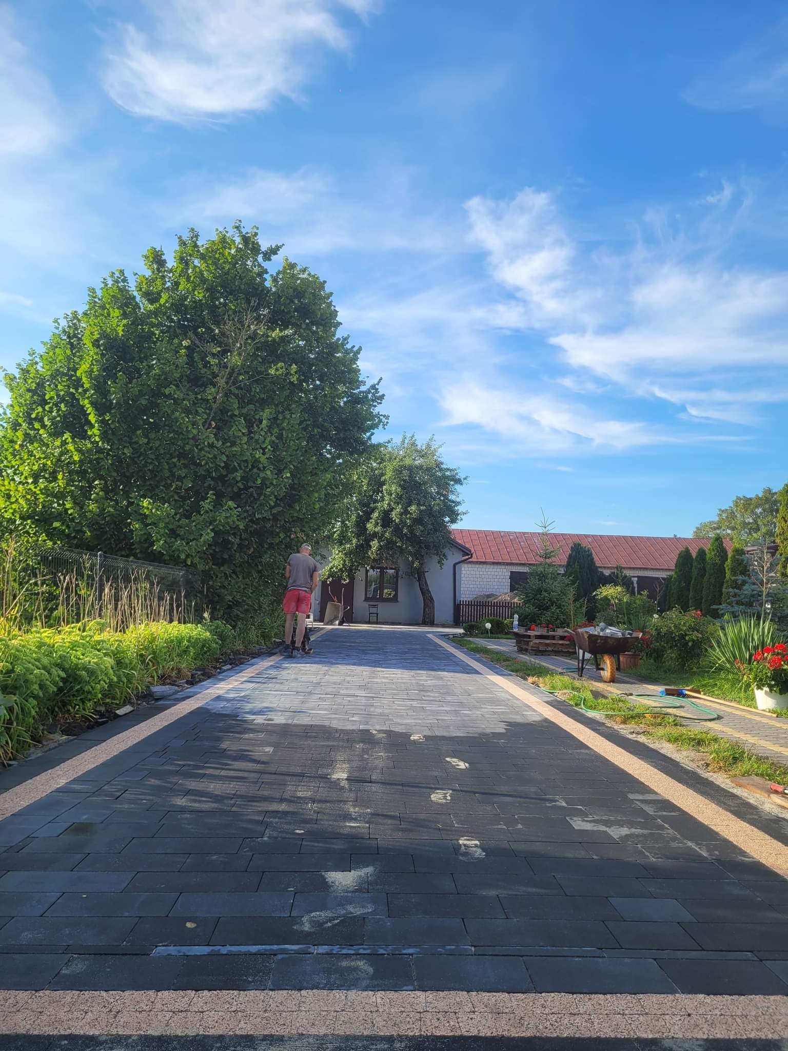 Świeżo ułożona, czarna kostka brukowa z jasnymi obrzeżami prowadząca do domu, widoczne ślady stóp, pracownik zagęszcza powierzchnię, błękitne niebo z chmurami w tle.