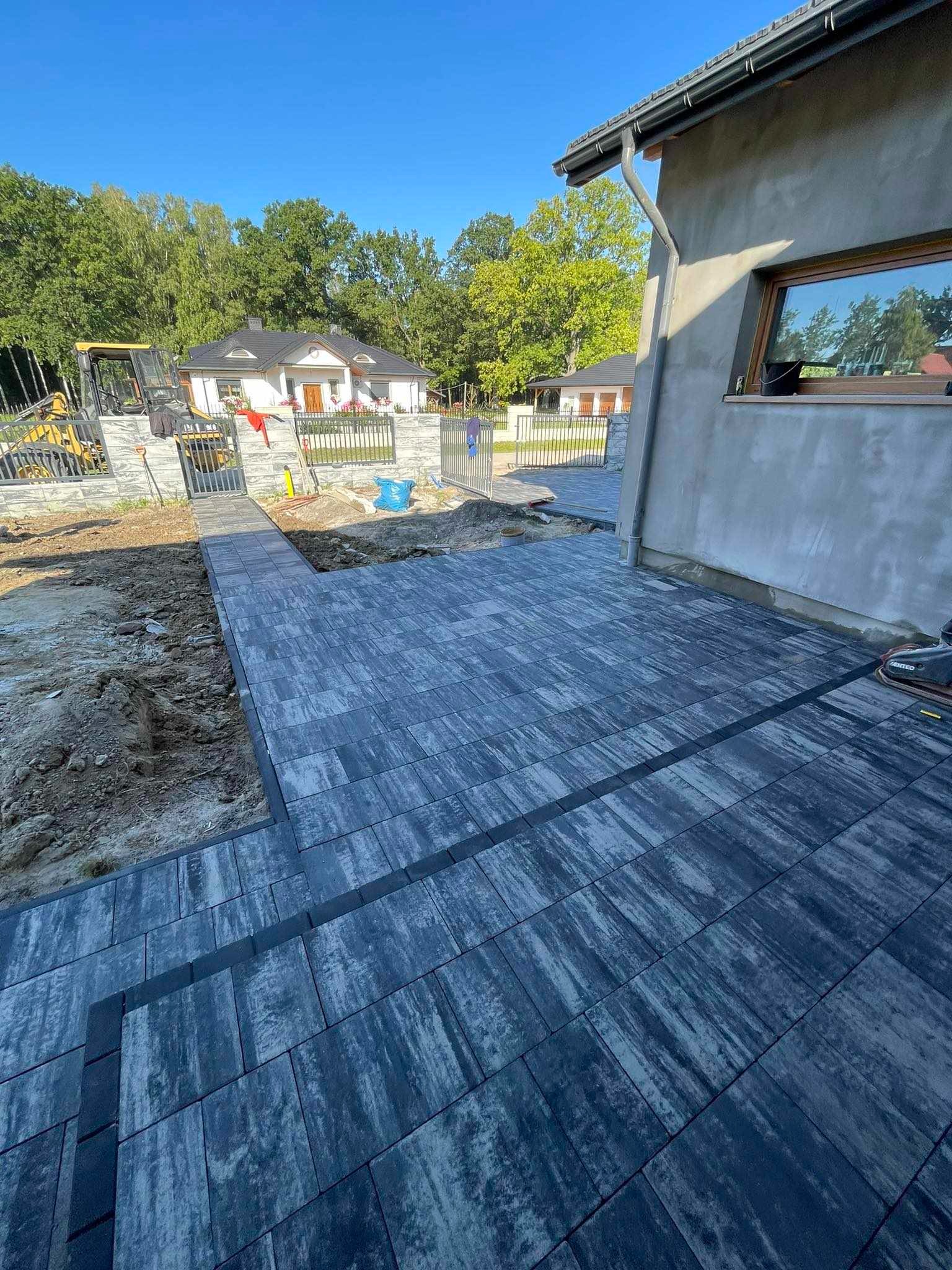 Ułożona szara kostka brukowa z czarnymi fugami tworząca taras i ścieżkę prowadzącą do furtki, w tle widoczny dom, trawnik i częściowo maszyna budowlana.