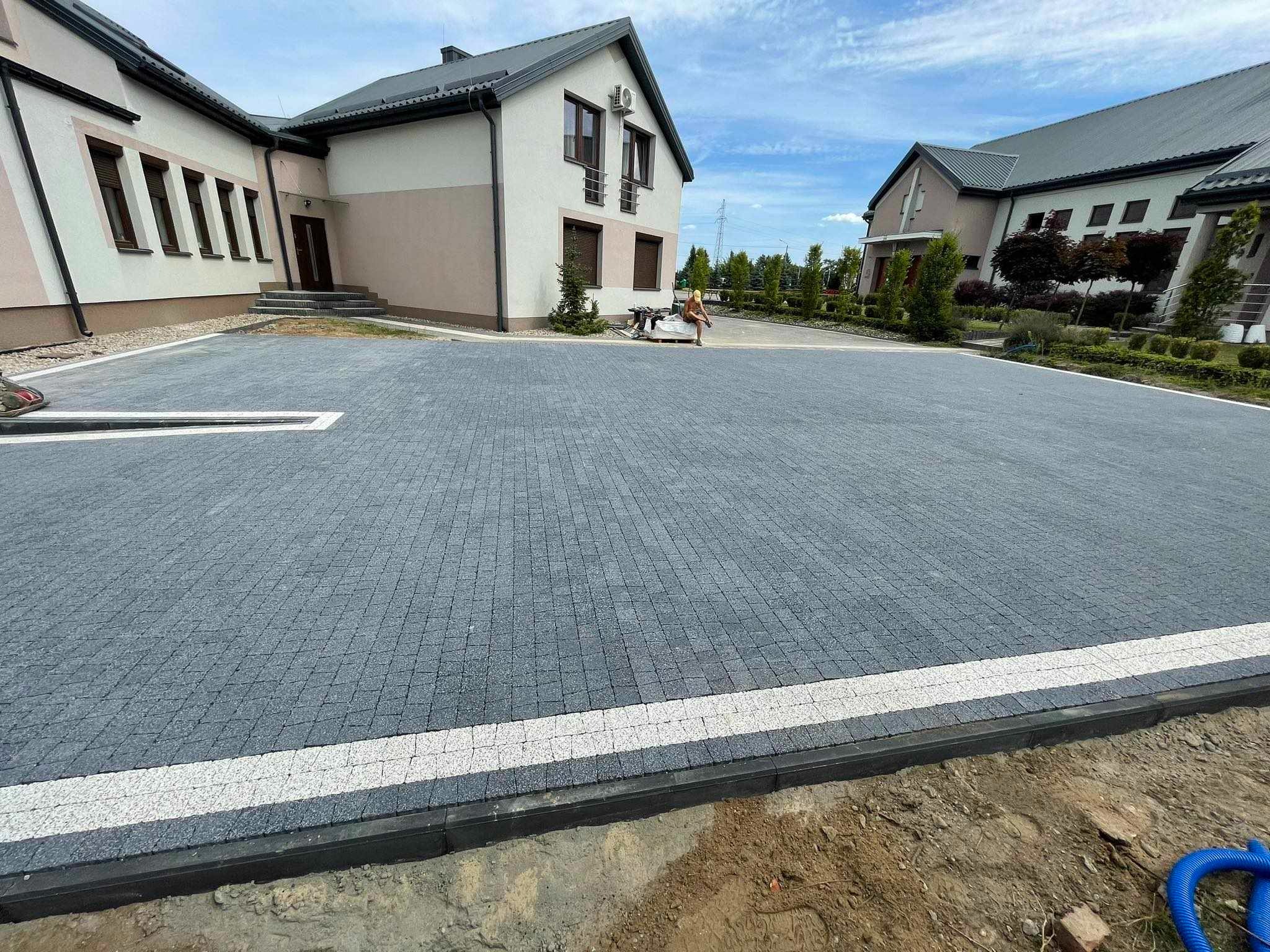 Nowa, duża powierzchnia z szarej kostki brukowej ułożonej we wzór jodełki, obramowana białymi pasami i czarnym obrzeżem, przed budynkiem z beżową elewacją i szarym dachem.