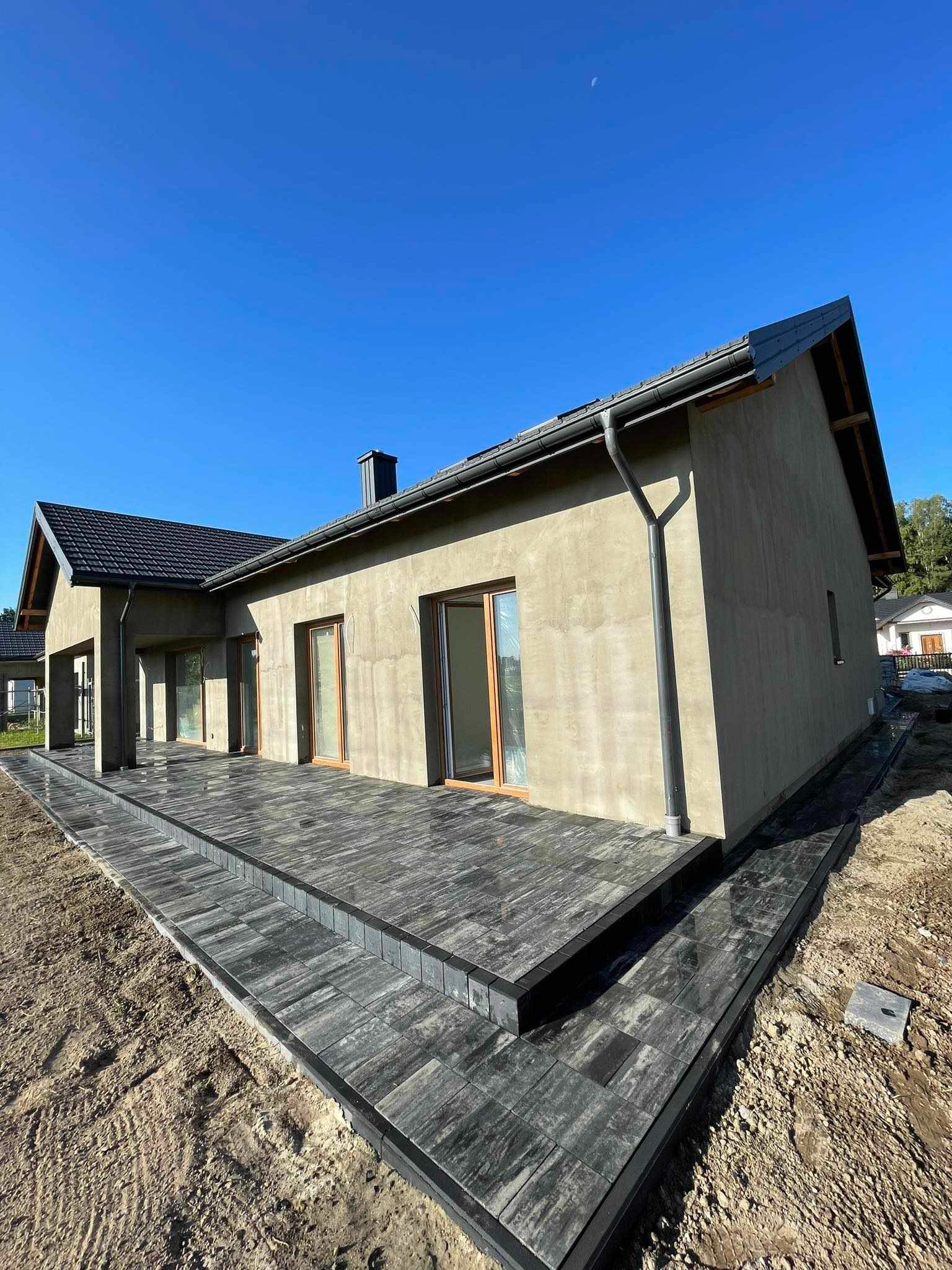 Nowoczesny dom jednorodzinny z wykończonym tarasem z szarej kostki brukowej, widok z niskiej perspektywy ukazujący detale ułożenia i kontrast z surową ziemią dookoła.