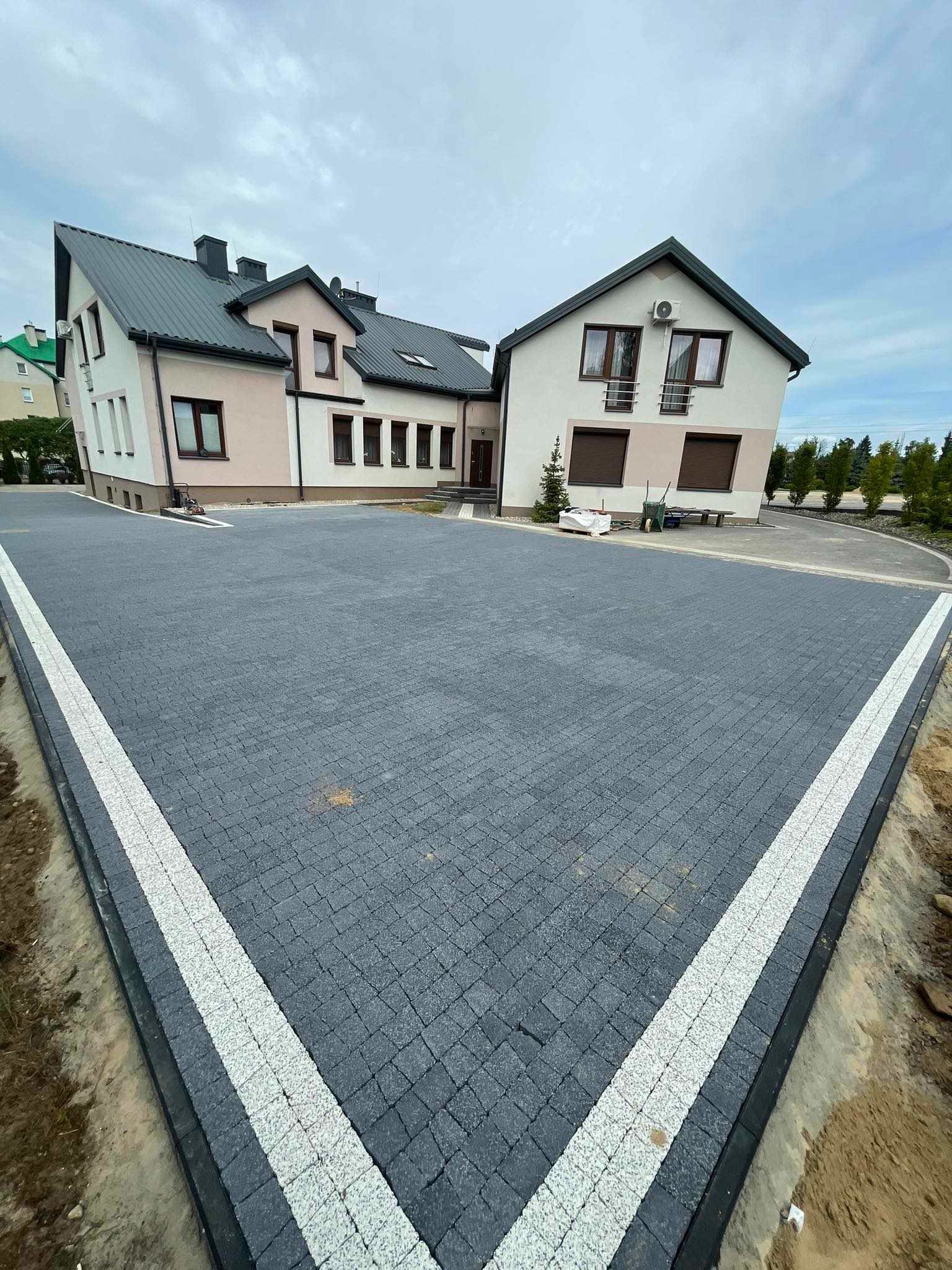Szeroki podjazd z ciemnoszarej kostki brukowej, obramowany białymi pasami, prowadzący do dwukondygnacyjnego budynku z jasną elewacją i ciemnym dachem.