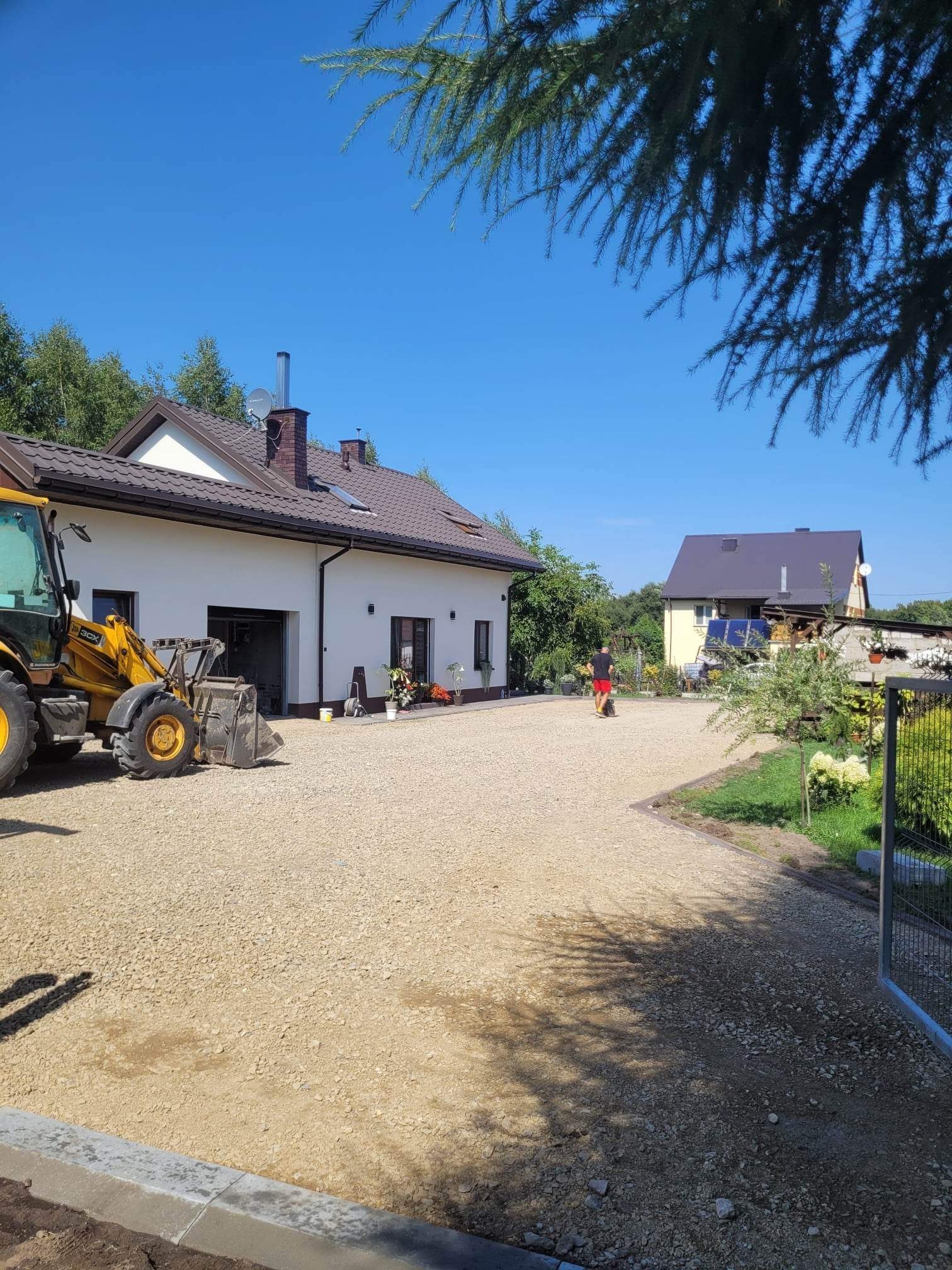Żółta ładowarka teleskopowa na żwirowym podłożu przed nowoczesnym domem z brązowym dachem, w tle widoczny drugi budynek i osoba spacerująca po żwirowym podwórku.