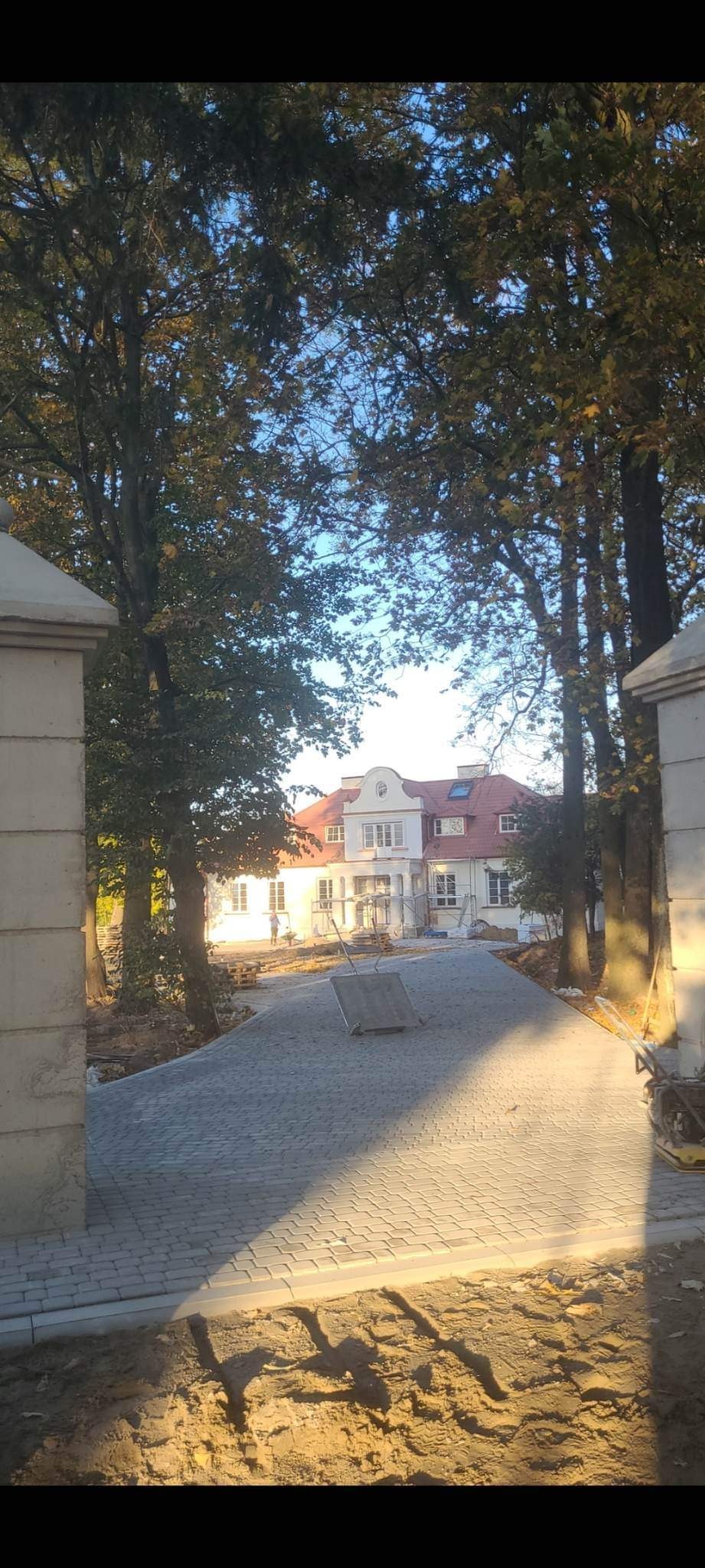 Nowo ułożona, szara kostka brukowa prowadząca do białego domu z czerwoną dachówką, widok przez kamienne filary bramy, częściowo zachmurzone niebo