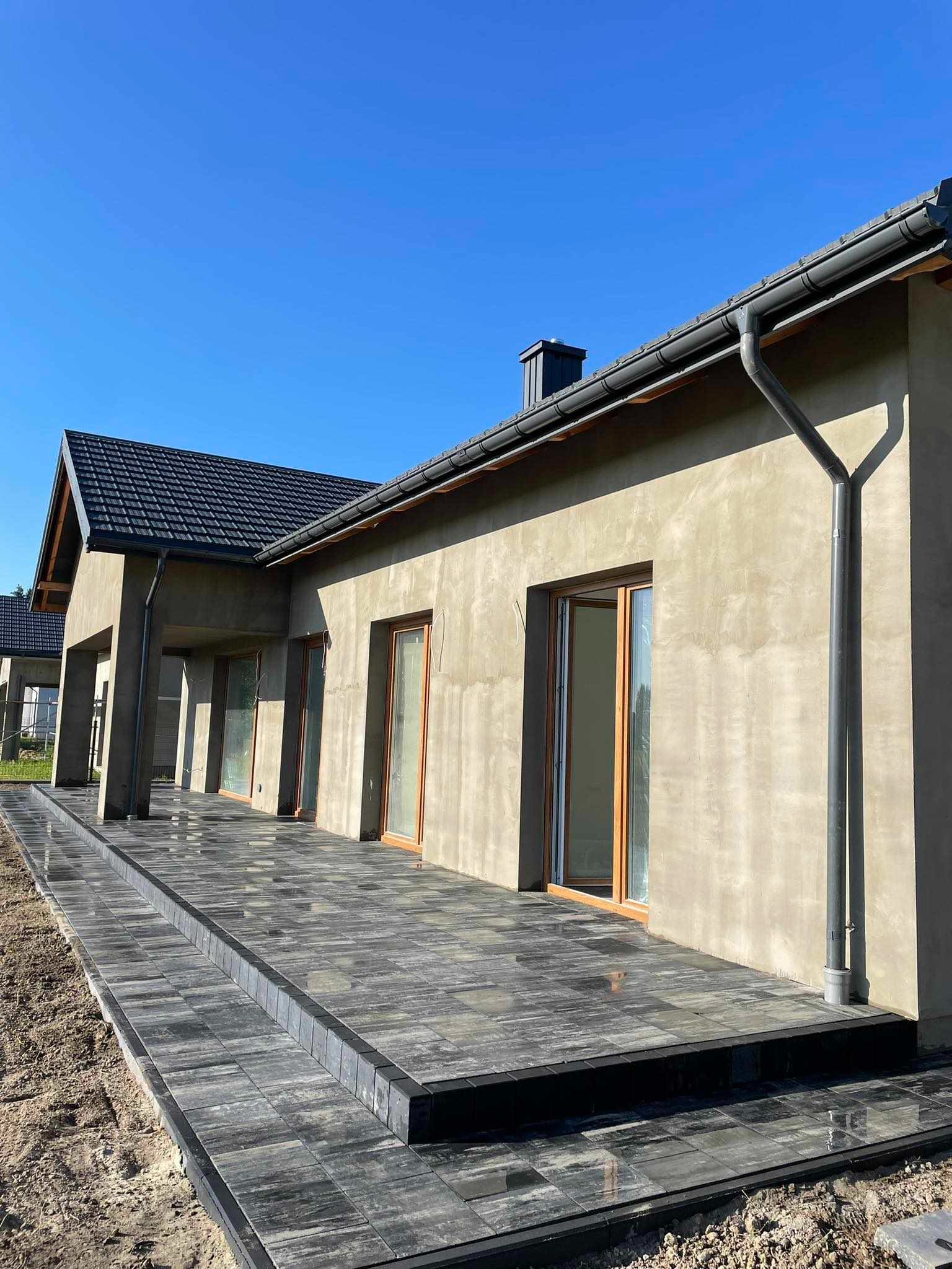 Ułożona, grafitowa kostka brukowa na tarasie domu jednorodzinnego z odkrytą elewacją, widoczne okna z drewnianą ramą i rynna.