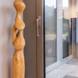 Szara rama okienna z widoczną fakturą drewna, obok drewniana rzeźba o organicznych kształtach na tle jasnej ściany.