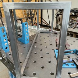 Maciej Szostakowski - Rama z profili stalowych spawana na stole spawalniczym z niebieskimi uchwytami, widoczne ślady po spawaniu i szlifowaniu, w tle inne konstrukcje stalowe.