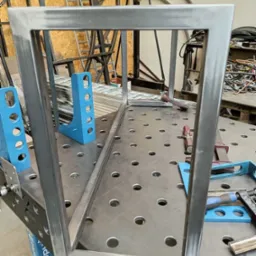 Rama z profili stalowych spawana na stole spawalniczym z niebieskimi uchwytami, widoczne ślady po spawaniu i szlifowaniu, w tle inne konstrukcje stalowe.