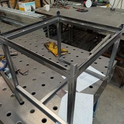 Maciej Szostakowski - Metalowa rama stołu spawana z profili na perforowanym stole warsztatowym, żółta rękawica robocza i plany konstrukcyjne obok, w tle narzędzia i spawacz przy pracy.