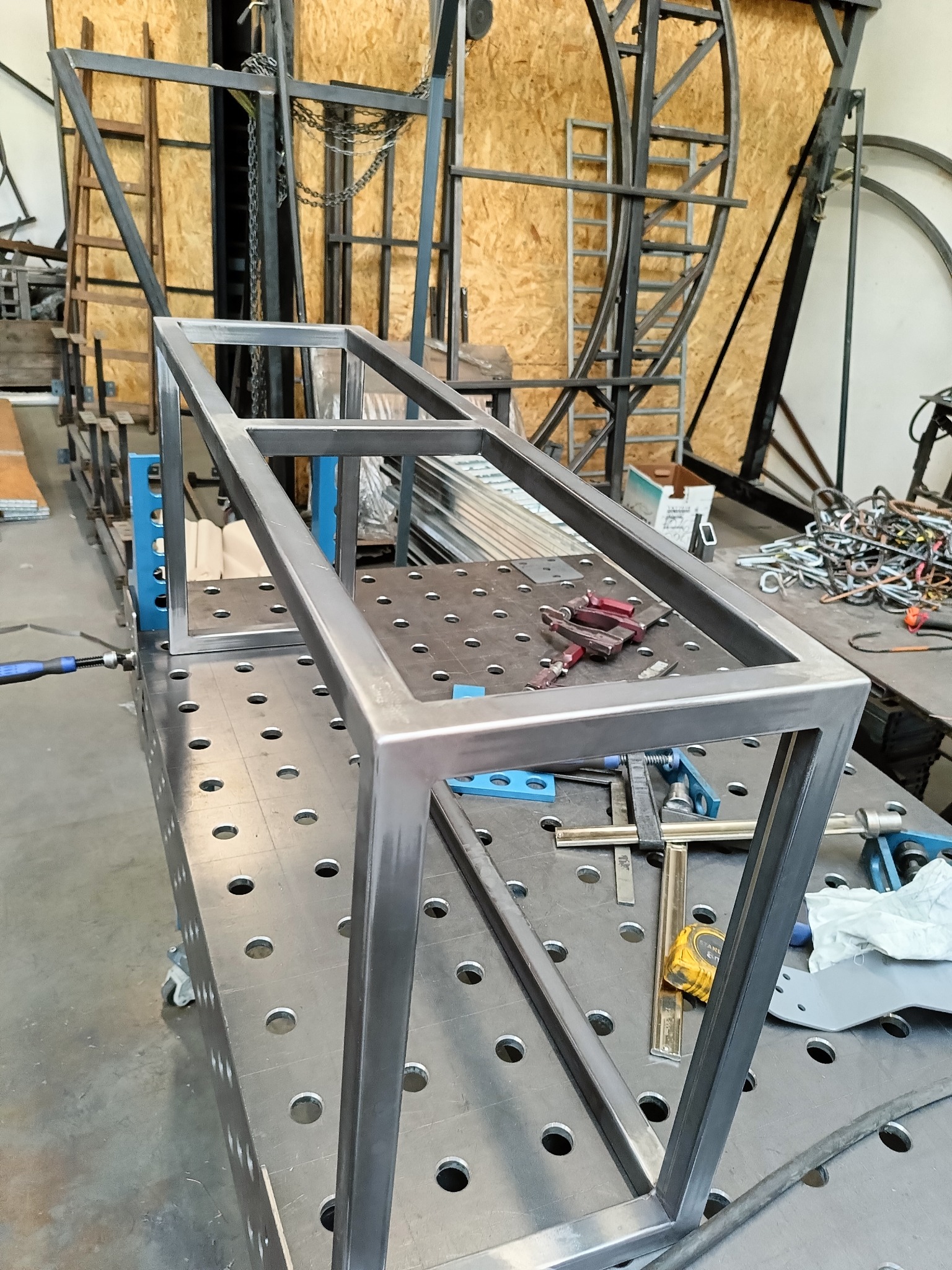 Stalowa rama spawana na perforowanym stole spawalniczym, narzędzia i akcesoria w tle, widoczne ślady obróbki metalu.