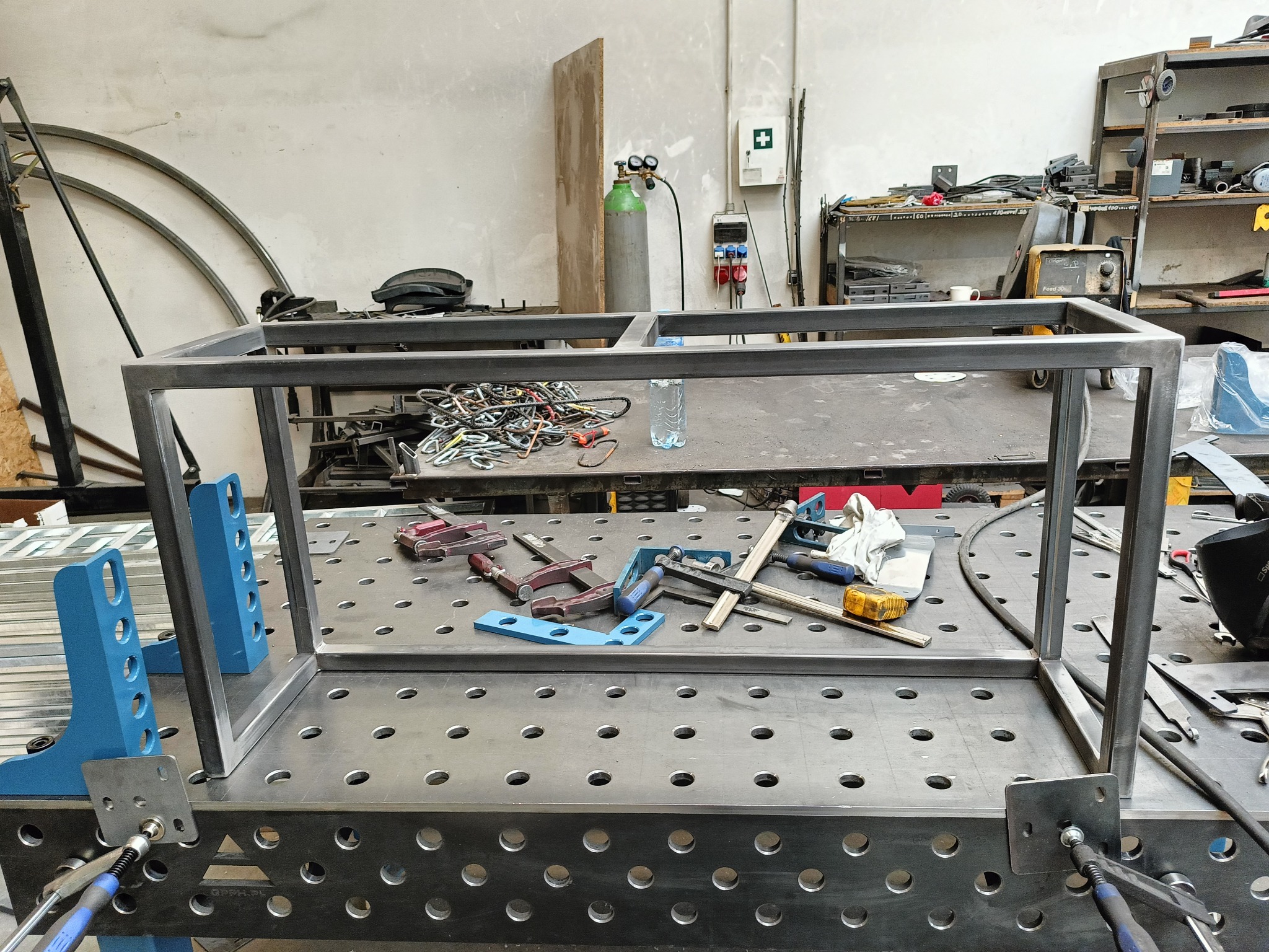 Stalowa rama meblowa w trakcie montażu na perforowanym stole spawalniczym, zabezpieczona niebieskimi zaciskami, z widocznymi narzędziami i butlą z gazem w tle.