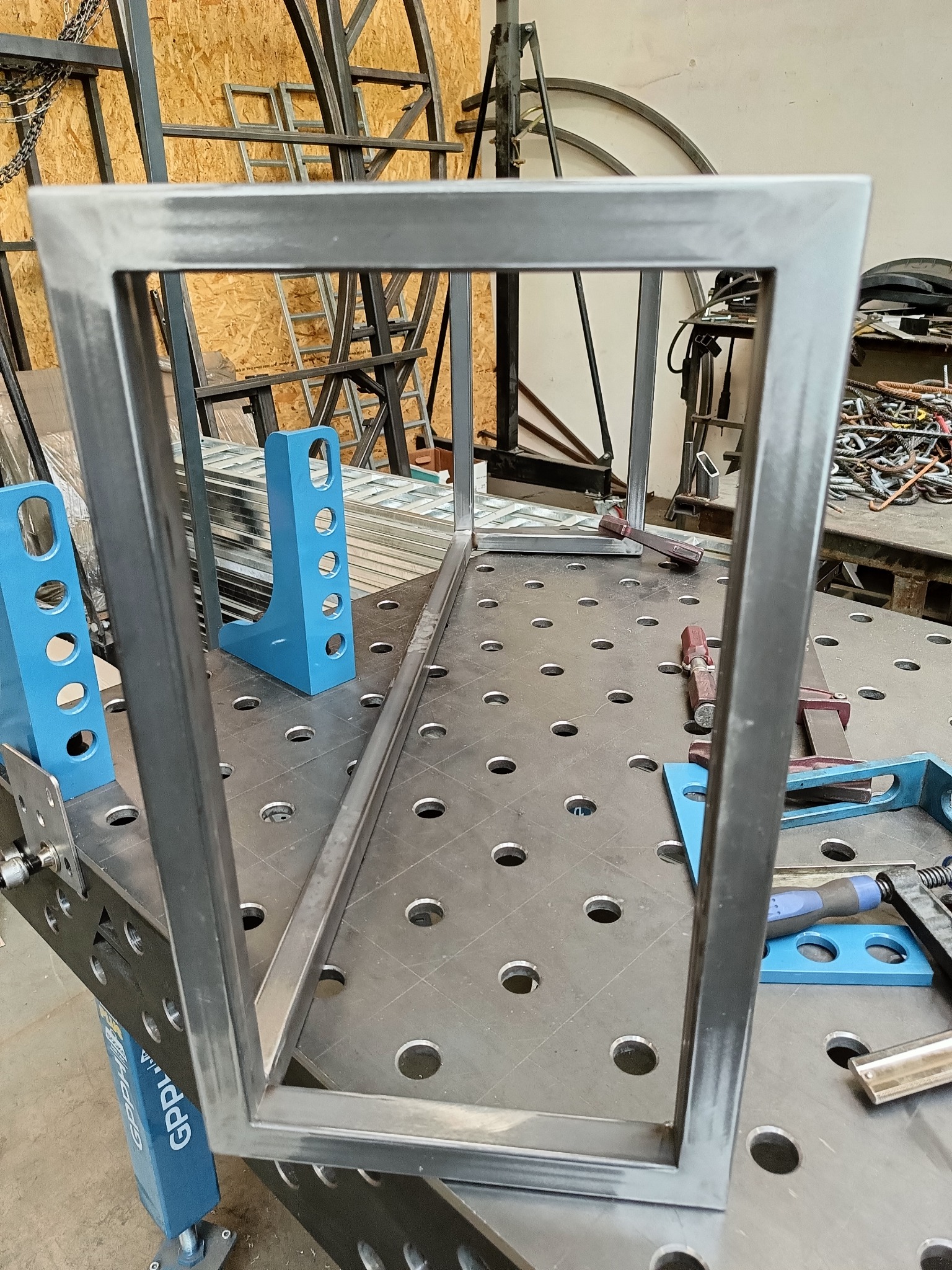 Rama z profili stalowych spawana na stole spawalniczym z niebieskimi uchwytami, widoczne ślady po spawaniu i szlifowaniu, w tle inne konstrukcje stalowe.