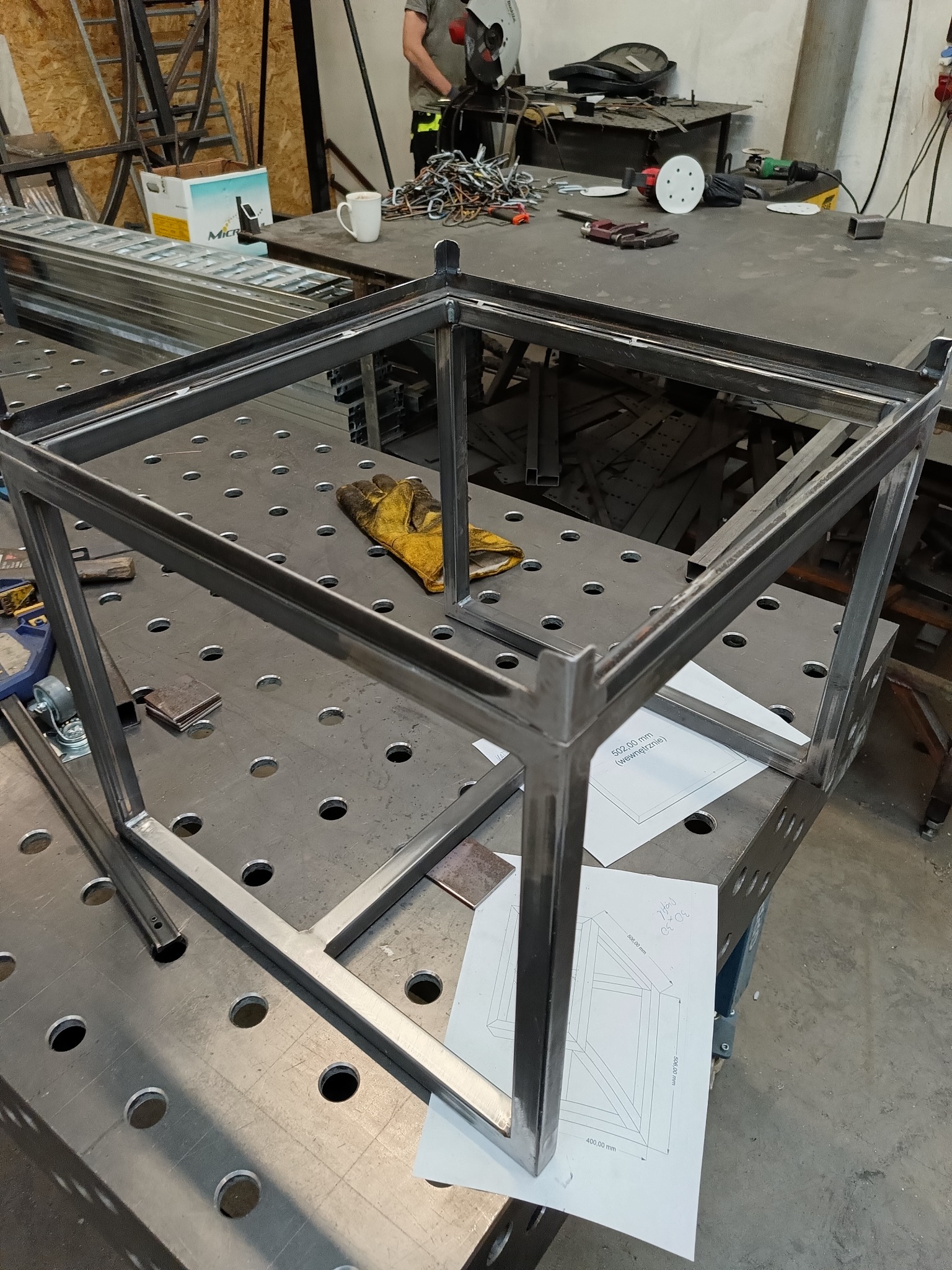 Metalowa rama stołu spawana z profili na perforowanym stole warsztatowym, żółta rękawica robocza i plany konstrukcyjne obok, w tle narzędzia i spawacz przy pracy.