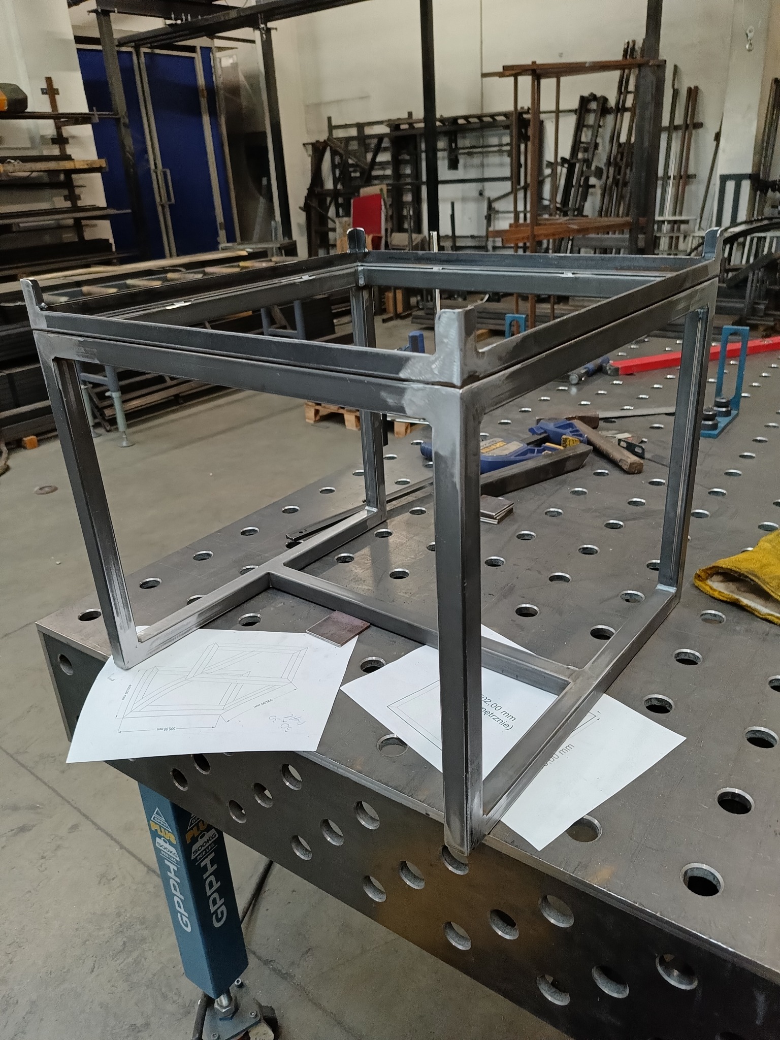 Metalowa konstrukcja szkieletowa, prawdopodobnie stelaż meblowy, spawana w Koleczkowie, ustawiona na perforowanym stole warsztatowym z widocznymi narzędziami i rysunkiem technicznym.