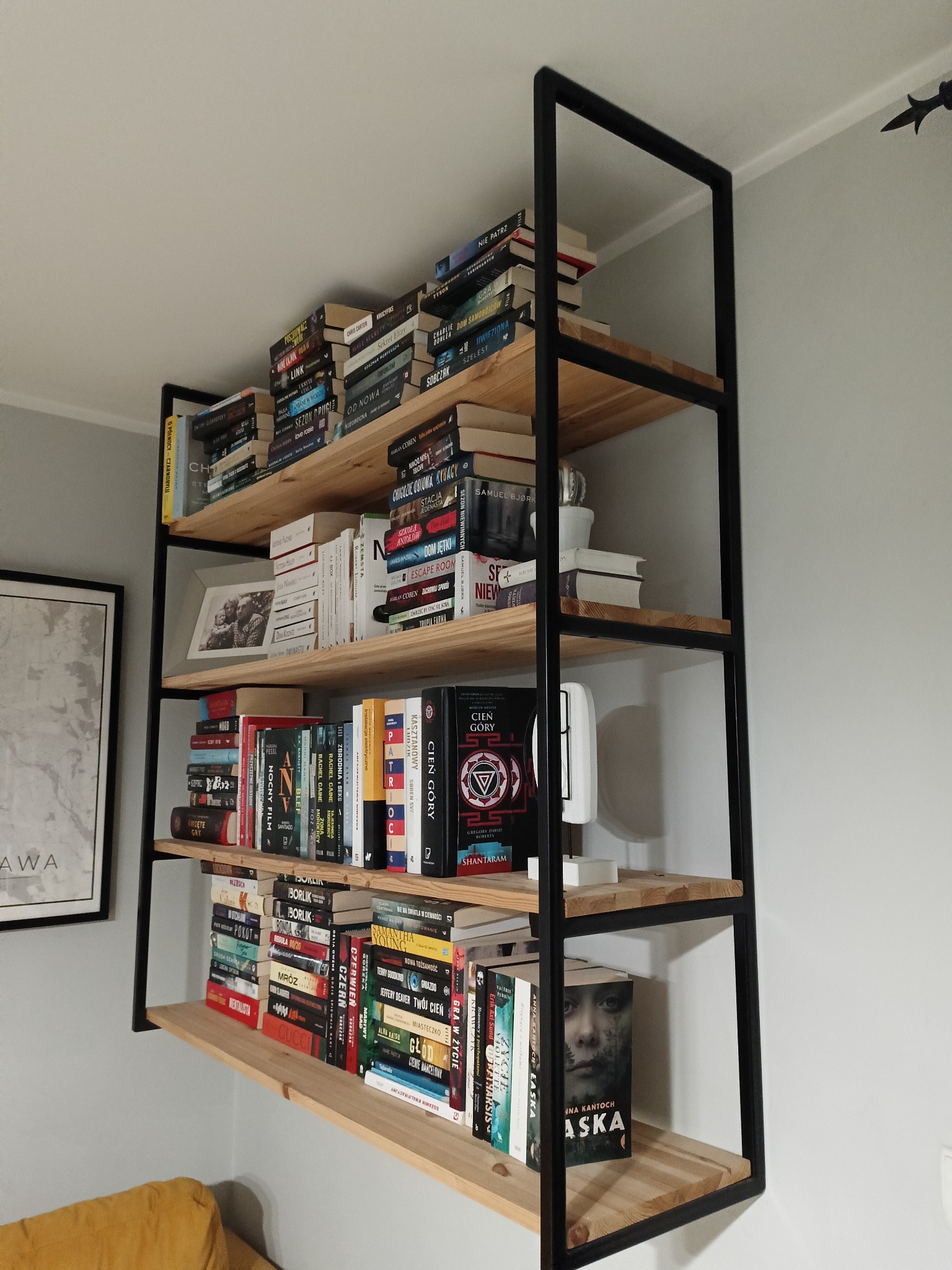 Ręcznie wykonany regał z metalową ramą i drewnianymi półkami, wypełniony książkami, ramką ze zdjęciem i dekoracjami, ustawiony przy szarej ścianie.