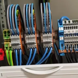 Wnętrze skrzynki elektrycznej z widocznymi przewodami w kolorach niebieskim, szarym i zielono-żółtym, podłączonymi do listew zaciskowych na szynach DIN.