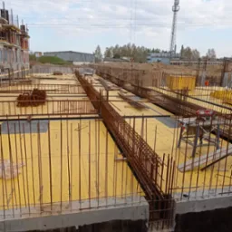 Zbrojenie fundamentów z żółtą szalunką i wystającymi prętami pionowymi, widok na plac budowy z wieżą telekomunikacyjną w tle.