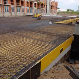 Zbrojenie płyty fundamentowej budynku w trakcie budowy, widoczny szkielet z prętów stalowych na żółtym podkładzie, w tle budynek w stanie surowym otwartym z czerwonej cegły i żółty agregat...