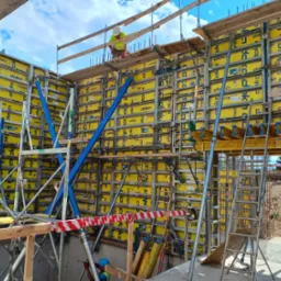 Konstrukcja szalunkowa Doka z żółtymi panelami, wzmocniona niebieskimi stężeniami, widoczny pracownik w kasku i kamizelce na górze, przygotowania do wylewania betonu.