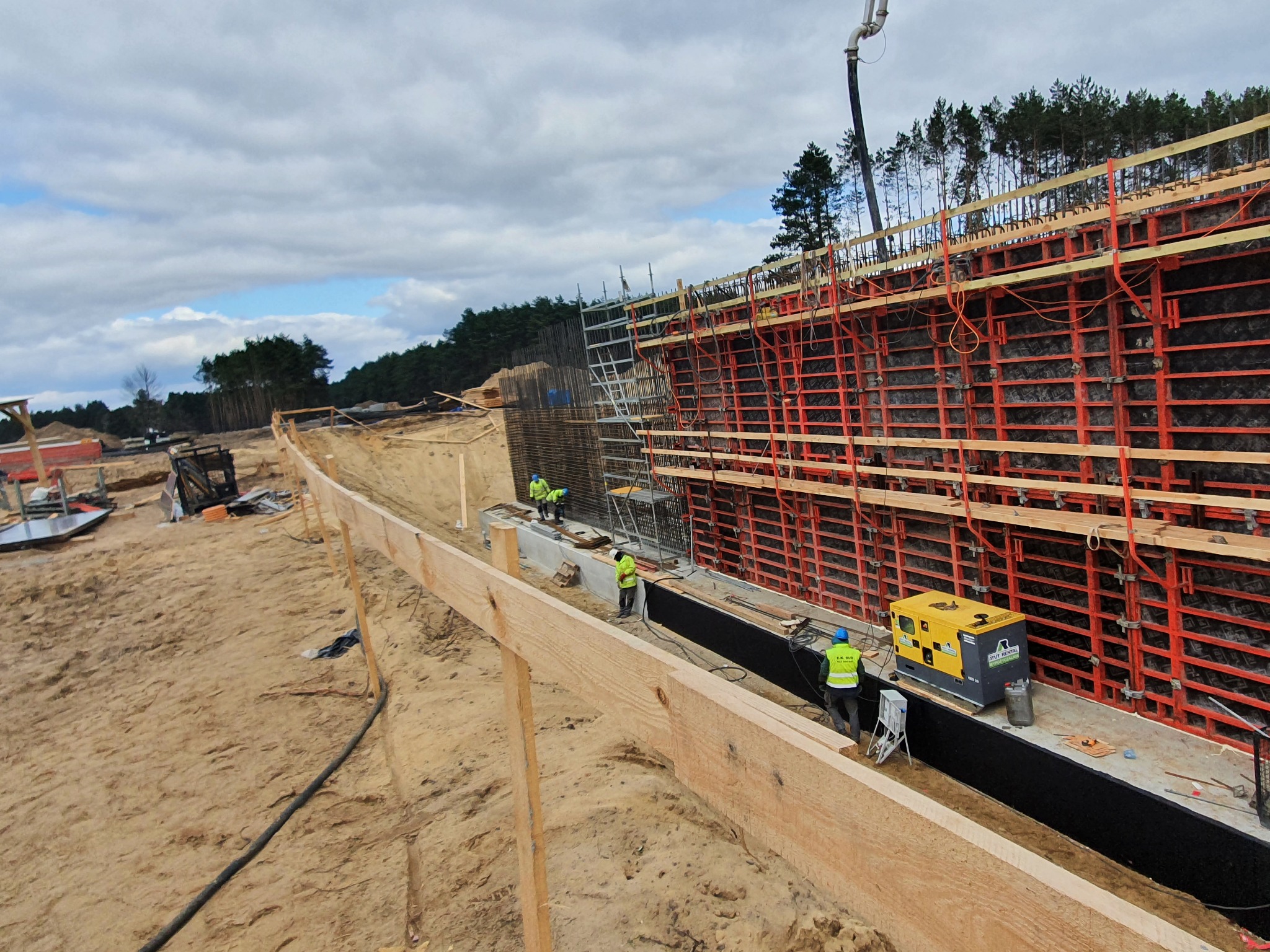 Budowa ściany oporowej z szalunkiem systemowym i zbrojeniem, widoczny generator prądu i pracownicy w odblaskowych kamizelkach na placu budowy.