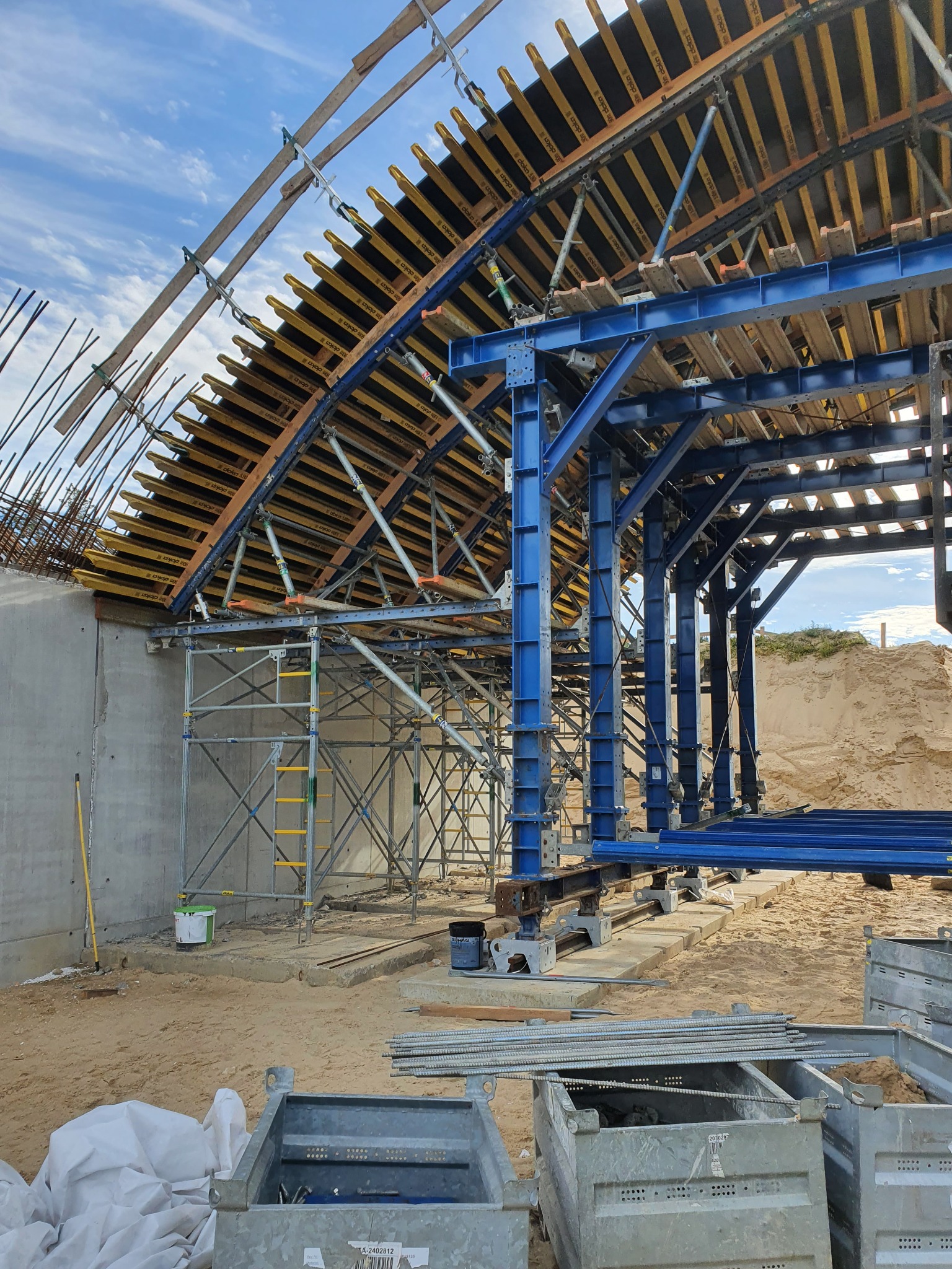 Konstrukcja szalunkowa z niebieskich stalowych belek i drewnianych paneli na budowie, widoczne rusztowania i betonowy mur w tle, ziemia piaszczysta.