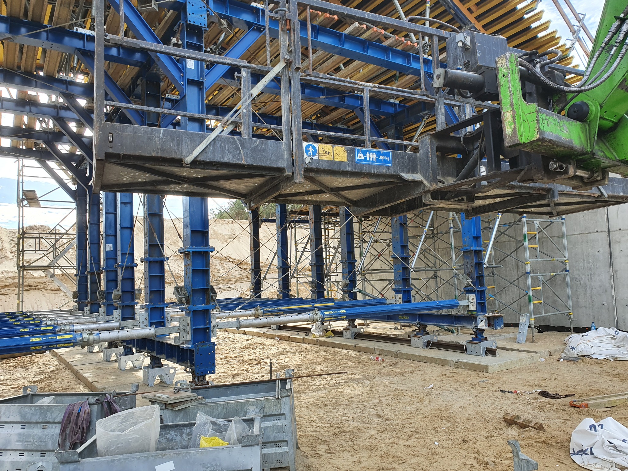 Szare szalunki ustawione na piasku, wsparte niebieskimi podporami, z widocznymi elementami dźwigu i szarym betonowym murem w tle.