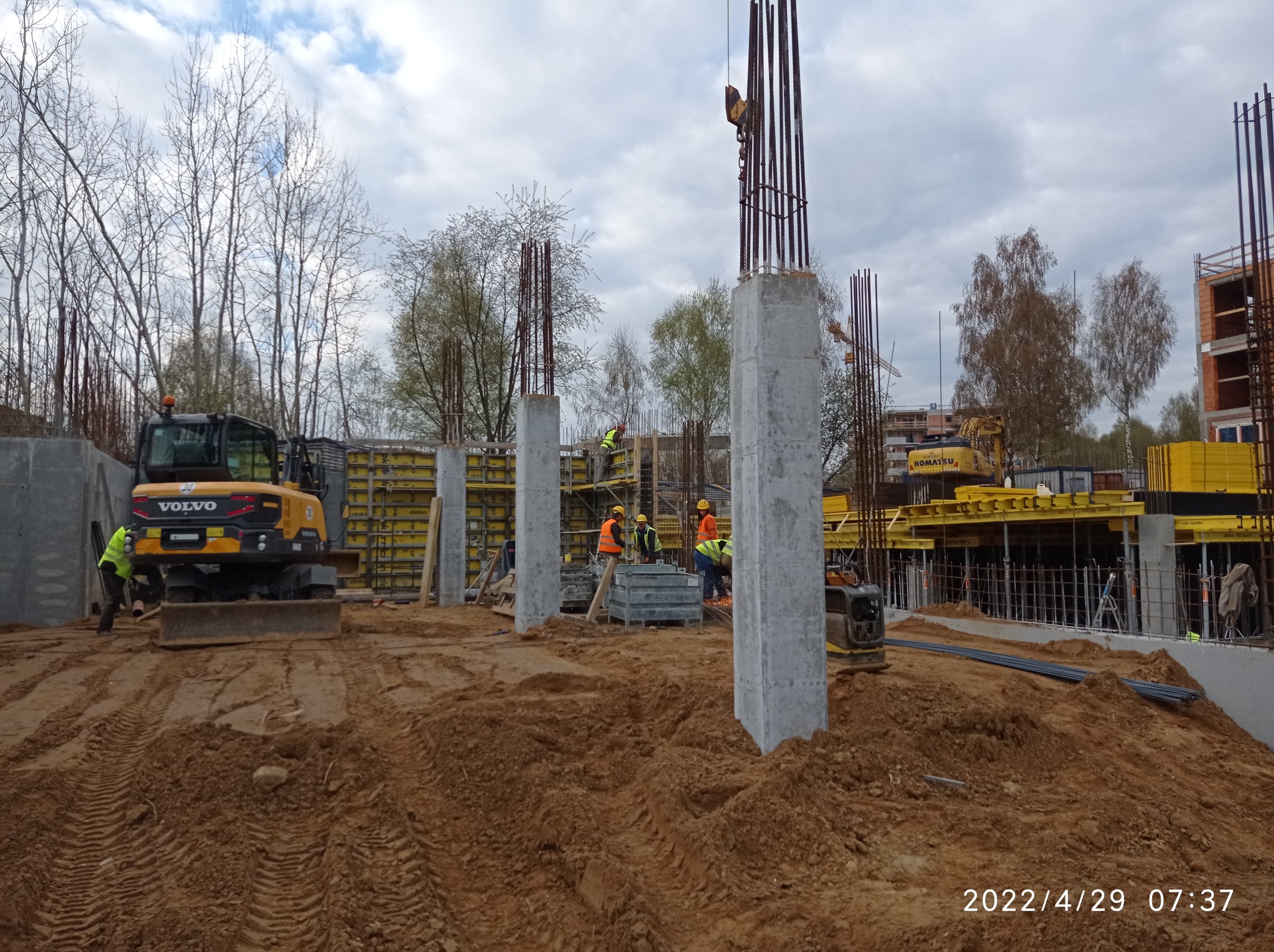 Budowa budynku żelbetowego: widok na plac budowy z fundamentami kolumn, zbrojeniami i pracującymi robotnikami w kaskach i kamizelkach odblaskowych, w tle żółty Volvo na nierównym terenie.
