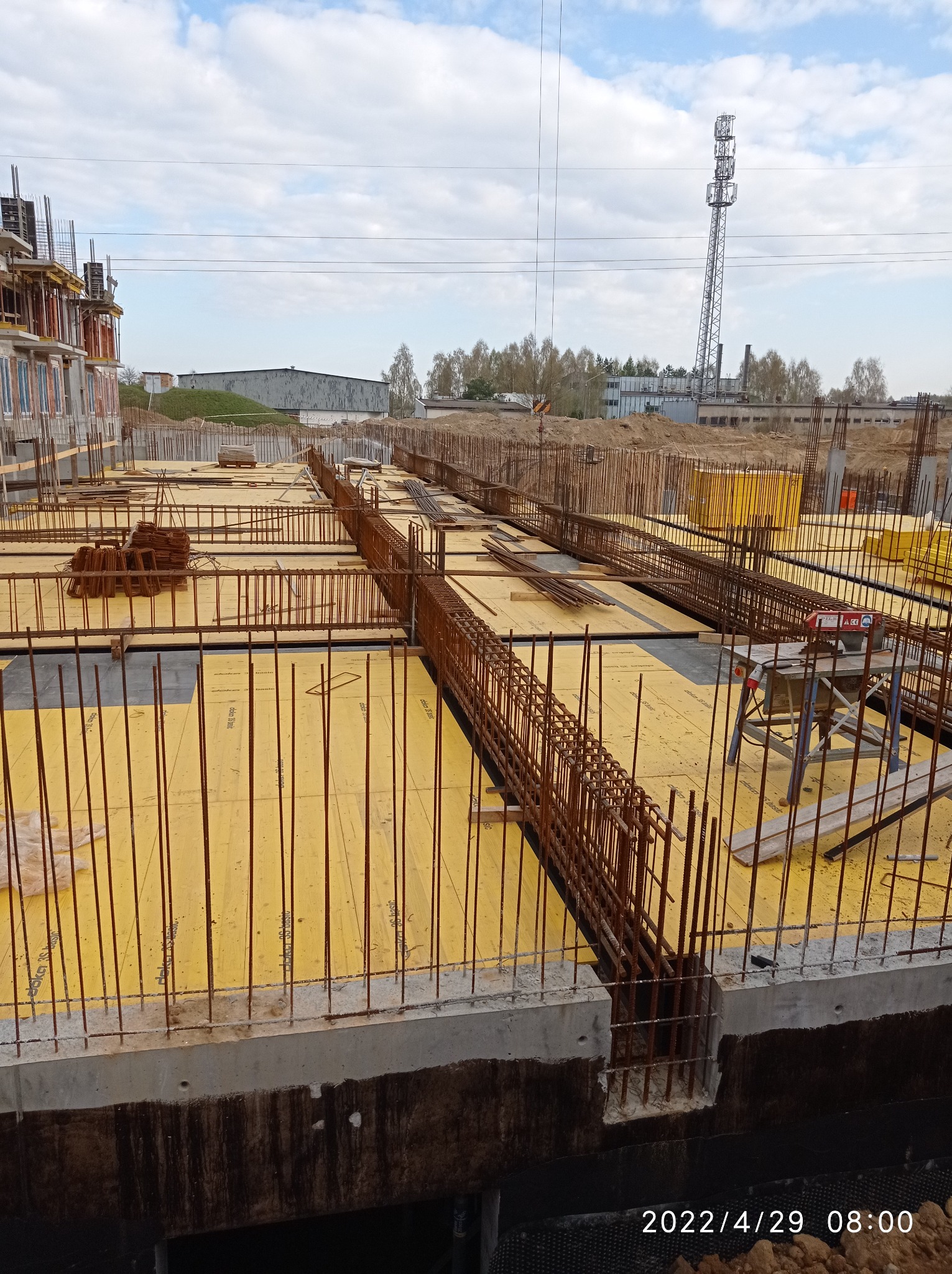 Zbrojenie fundamentów z żółtą szalunką i wystającymi prętami pionowymi, widok na plac budowy z wieżą telekomunikacyjną w tle.