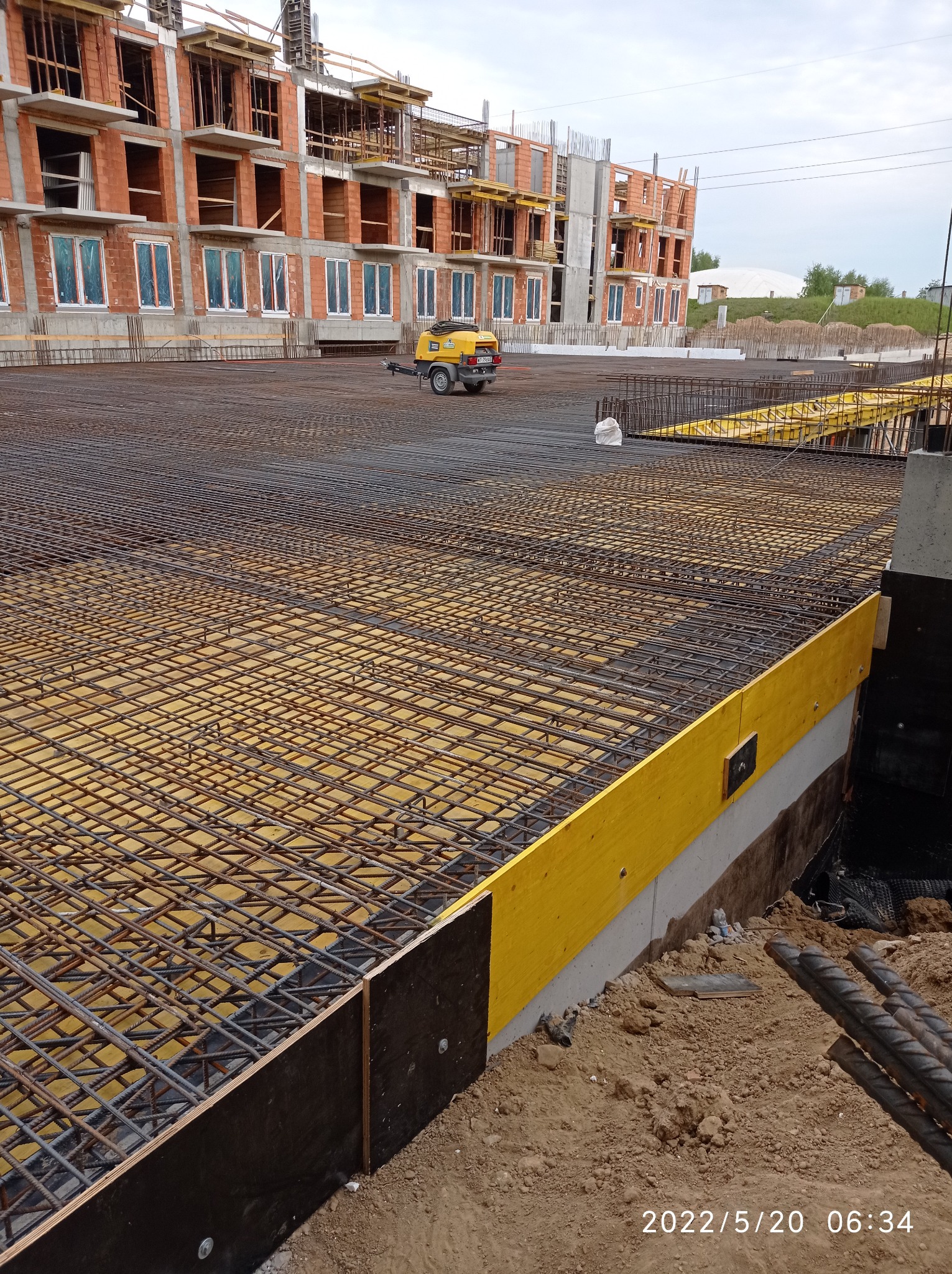 Zbrojenie płyty fundamentowej budynku w trakcie budowy, widoczny szkielet z prętów stalowych na żółtym podkładzie, w tle budynek w stanie surowym otwartym z czerwonej cegły i żółty agregat...