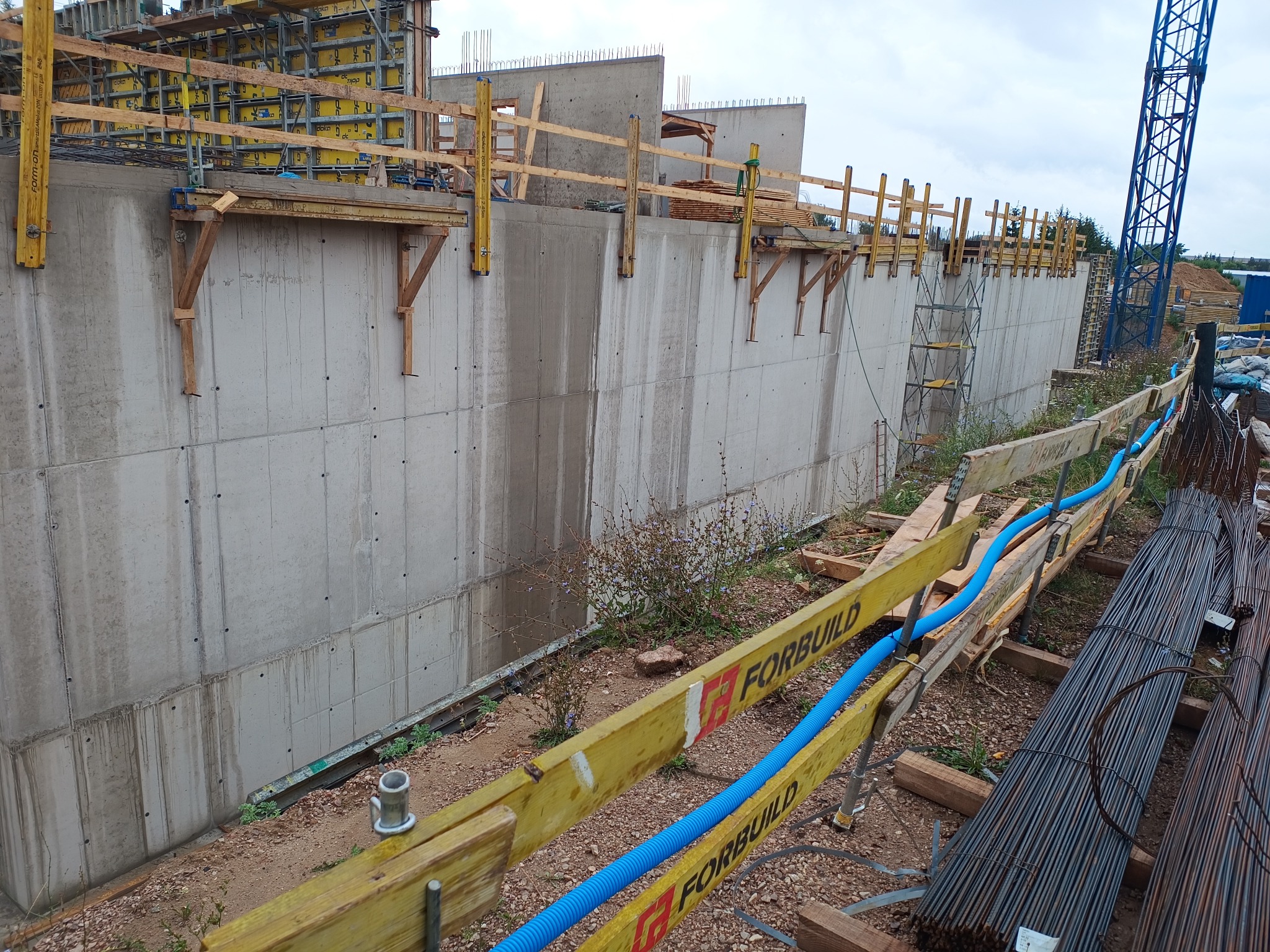 Betonowa ściana oporowa w trakcie budowy, z widocznymi szalunkami, deskami i niebieskimi rurami ochronnymi wzdłuż krawędzi, na tle placu budowy z żurawiem.
