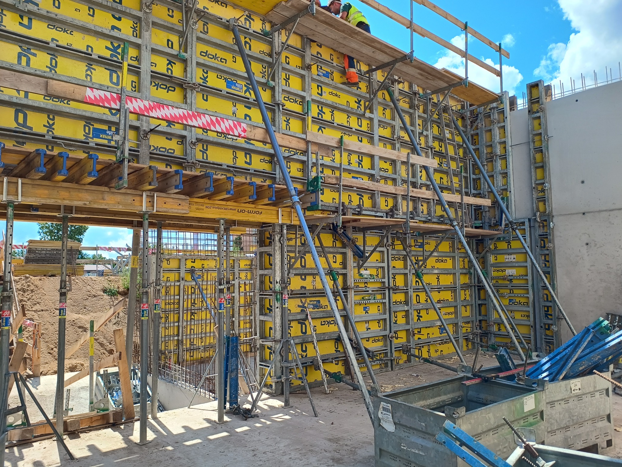 Szczegółowe ujęcie szalunków Doka wypełnionych żółtą pianką, przygotowanych do wylania betonu na budowie, z widocznymi elementami konstrukcyjnymi i pracownikami na górze.