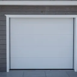 Zamknięta biała brama segmentowa z poziomymi panelami, otoczona białą ramą, na tle szarej, drewnianej elewacji garażu.