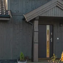 Elewacja domu z ciemnego drewna z nowoczesnymi oknami z roletami wewnętrznymi i drzwiami wejściowymi z ozdobnym panelem.